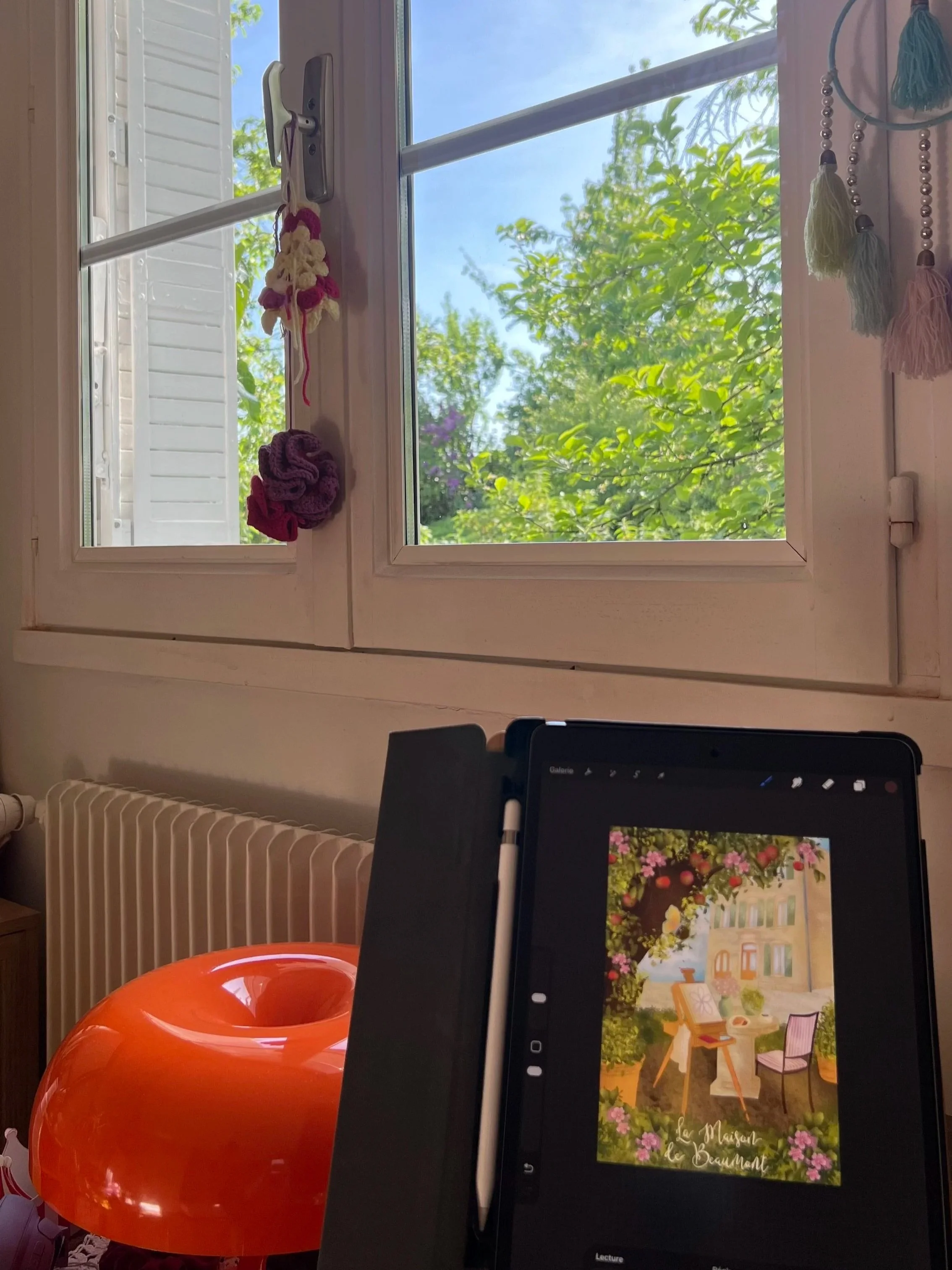 Open window with a view of green trees and a blue sky, decorated with hanging tassels and crocheted items, and a laptop displaying a garden scene.
