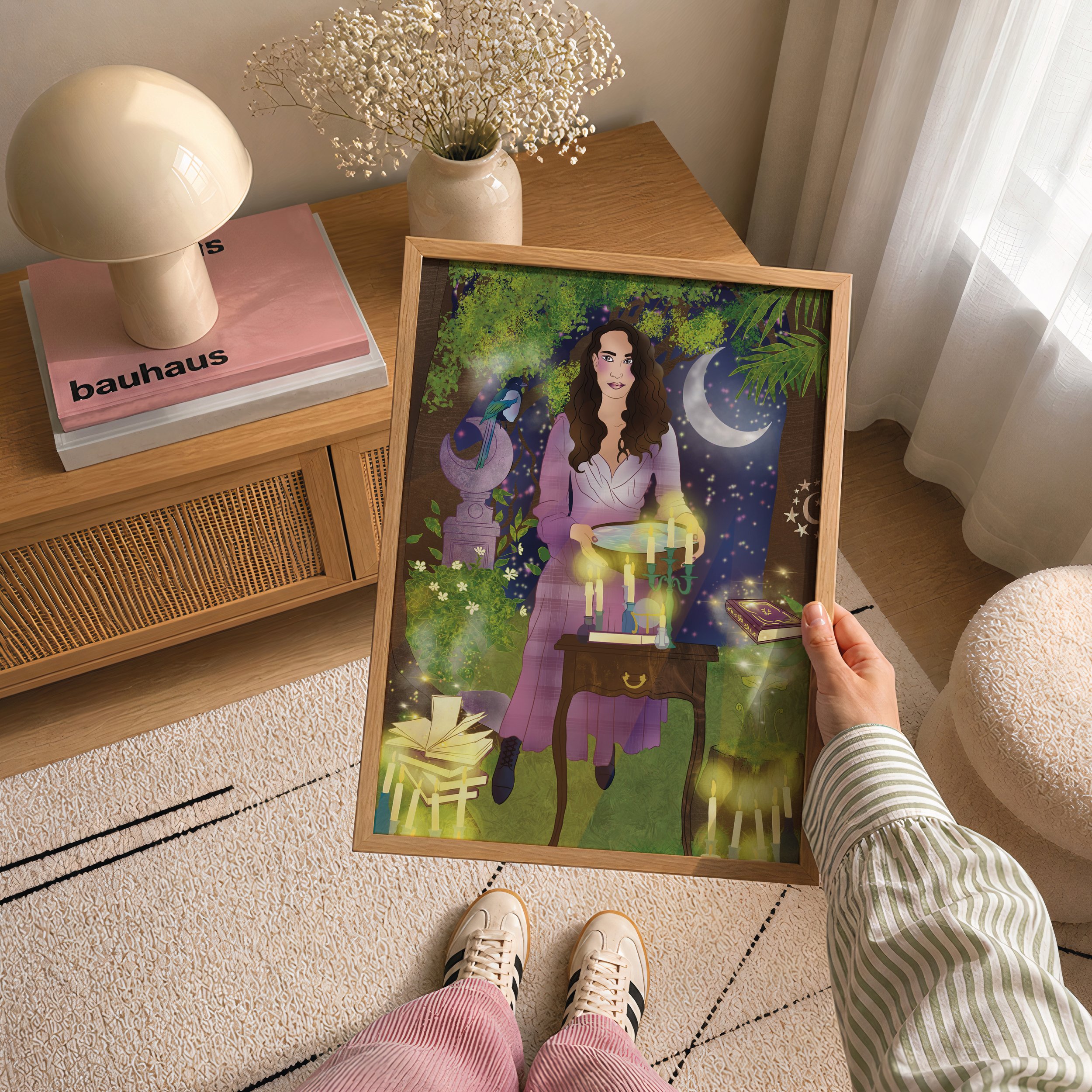 Top view of a wooden table with a white vase of dried flowers, a lamp, pink and white books, and a framed artwork featuring a woman with long brown hair, a moon, and lush green foliage, near a window with sheer curtains.