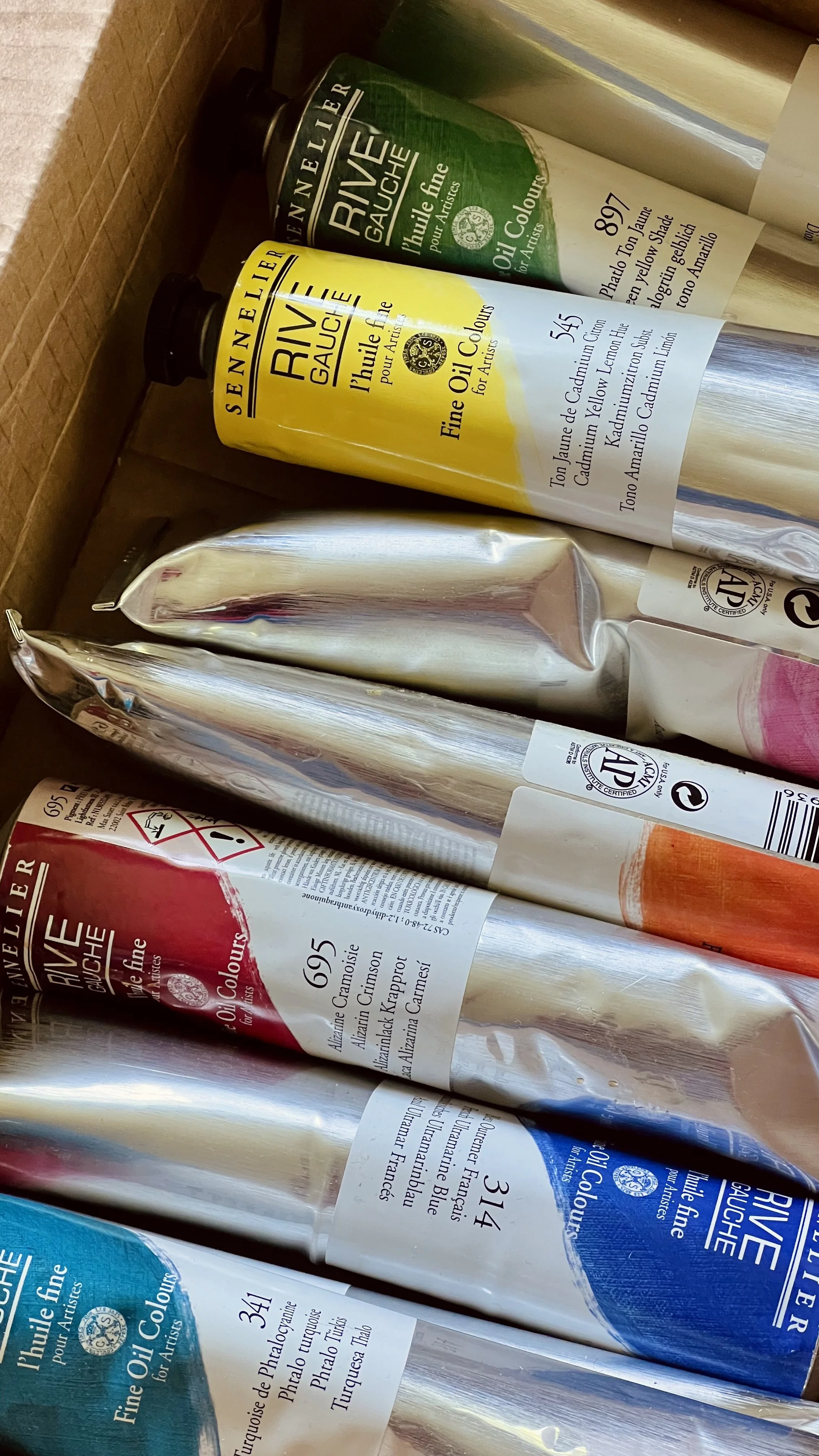 A close-up view of several tubes of oil paint in various colors including blue, red, yellow, green, and white, with labels showing brand and color information.