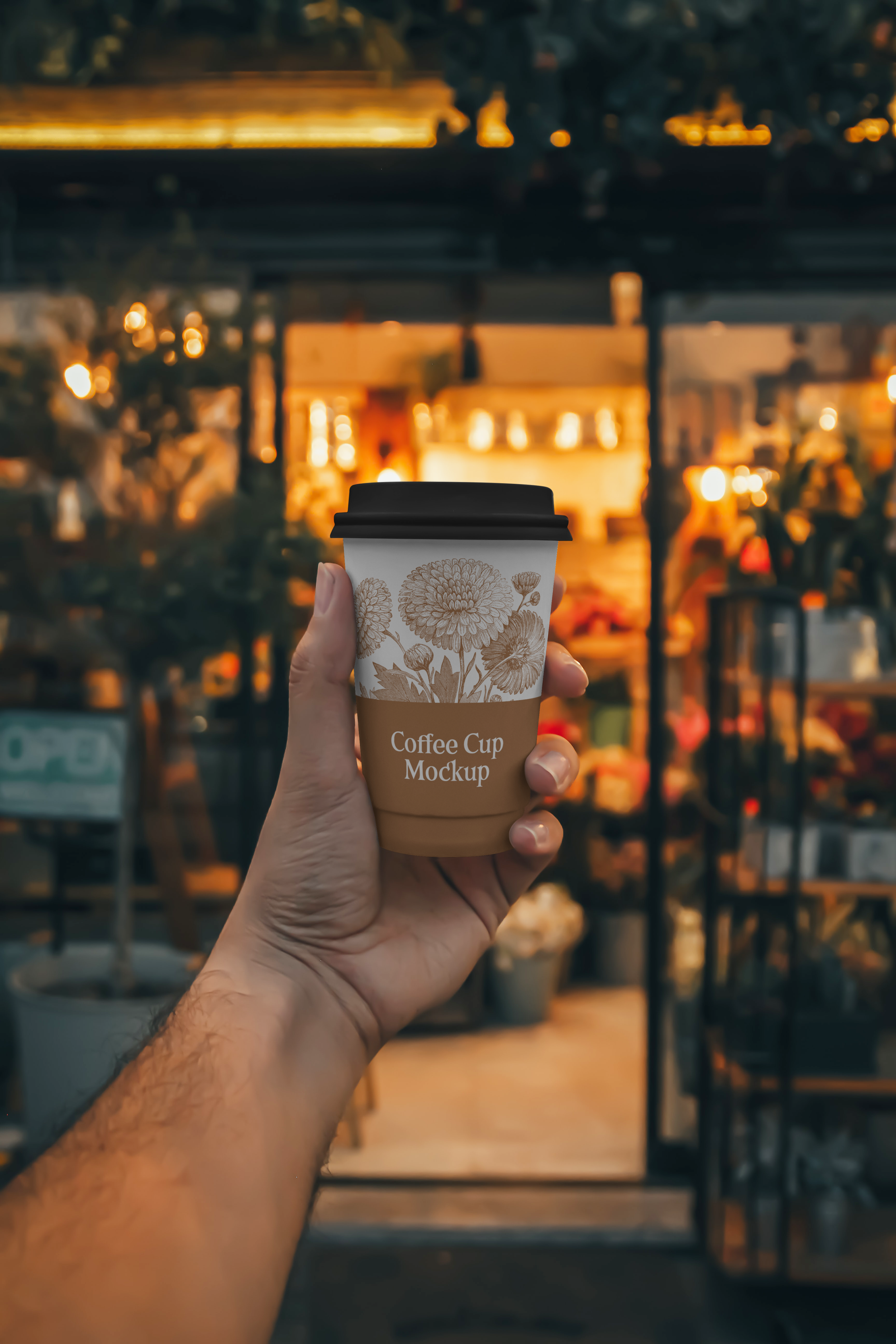 A hand holding a disposable coffee cup with a floral design and a black lid. The background features a cozy, warmly lit cafe with blurred lights and furniture.