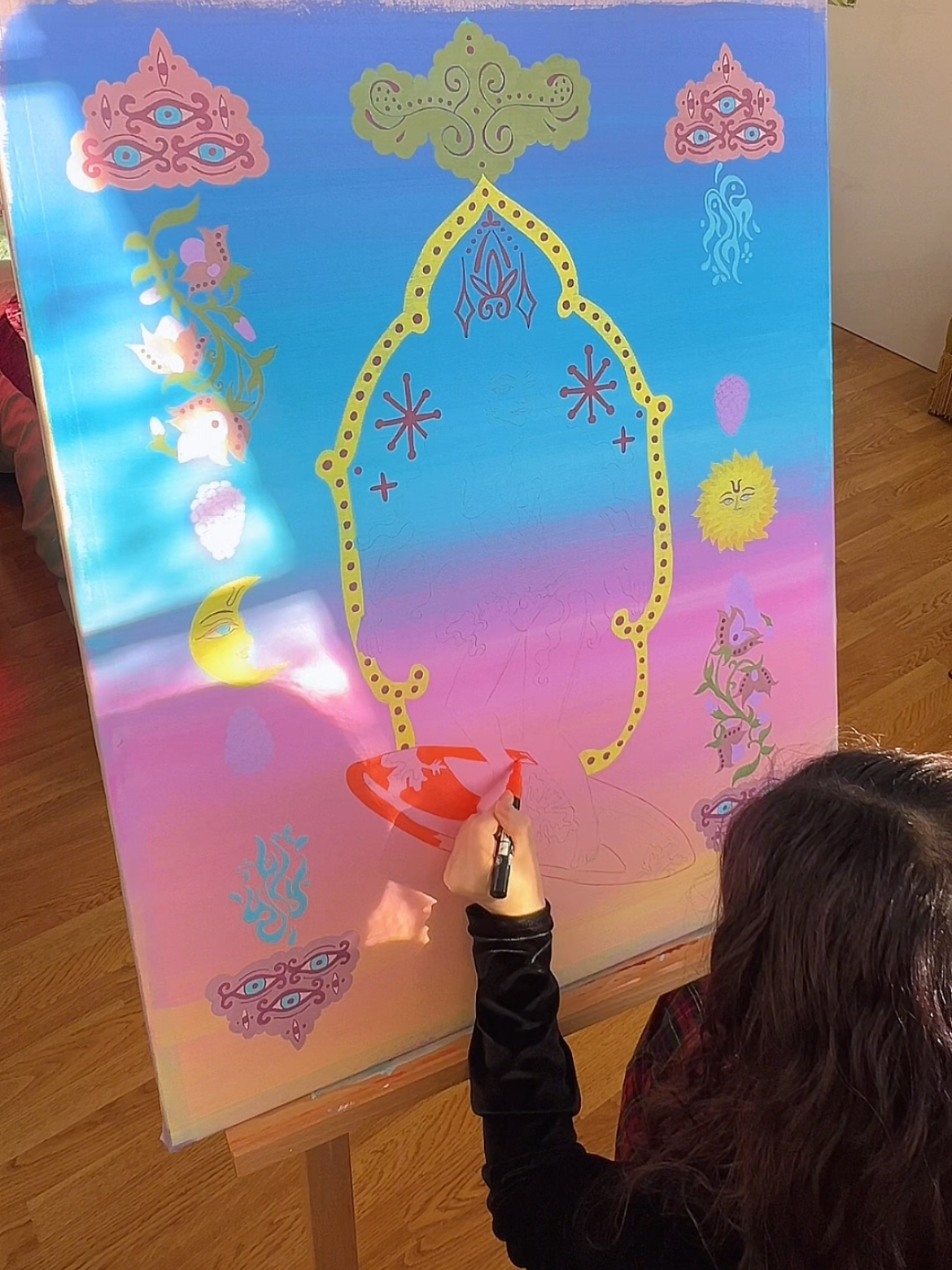 A person drawing colorful, fantasy-themed artwork on a large surface, which includes various mythical and celestial symbols such as moons, stars, eyes, a sun, and other abstract designs, with a gradient background of pink, blue, and purple.