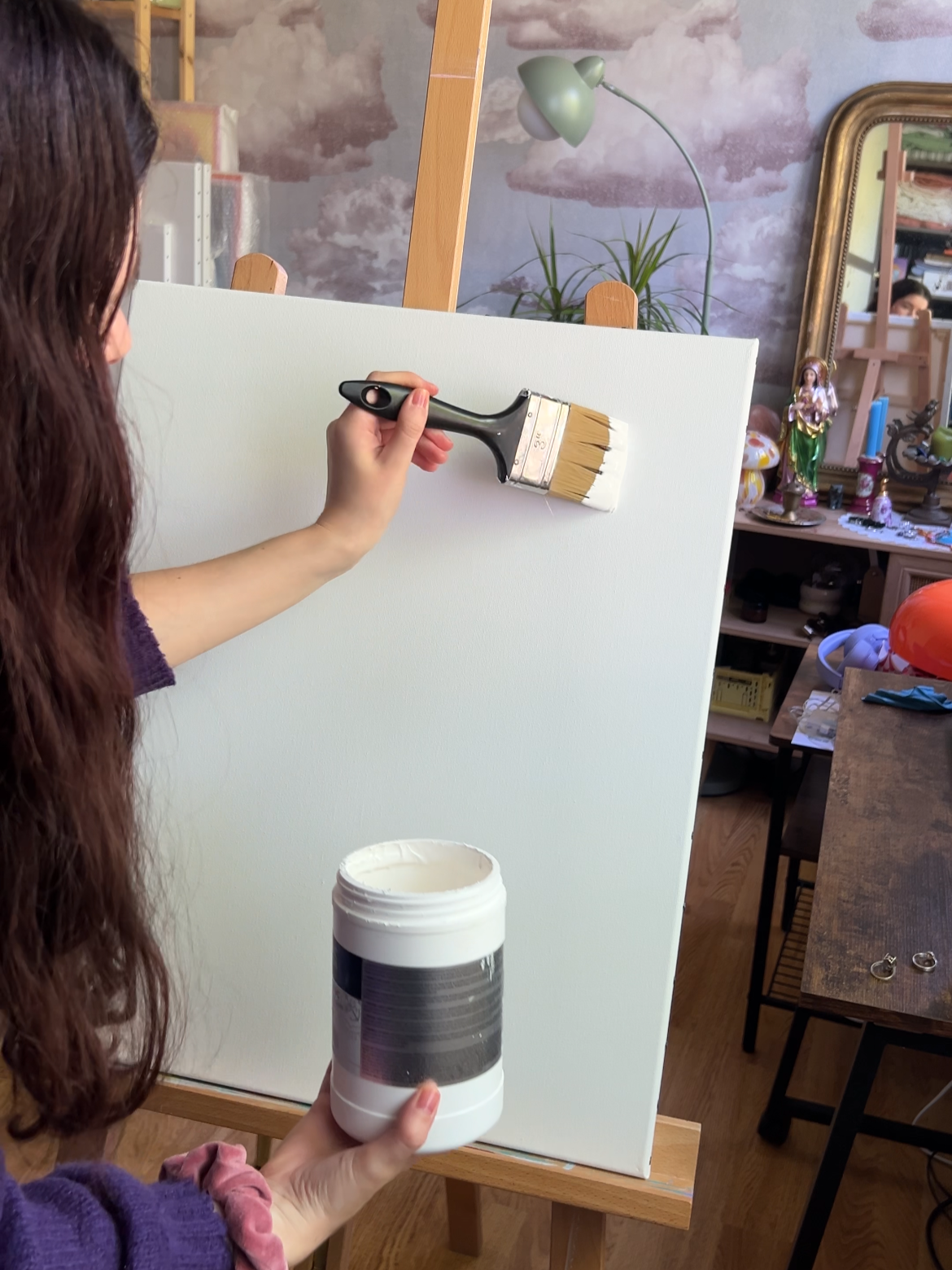 Person painting a blank white canvas with a paintbrush in a room filled with art supplies and decorations.