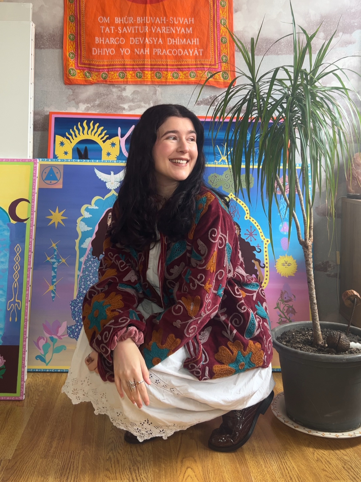 Young woman smiling, sitting on the floor near a potted plant, wearing a colorful embroidered jacket and a white dress with eyelet details, with colorful decorative posters and a tapestry on the wall behind her.