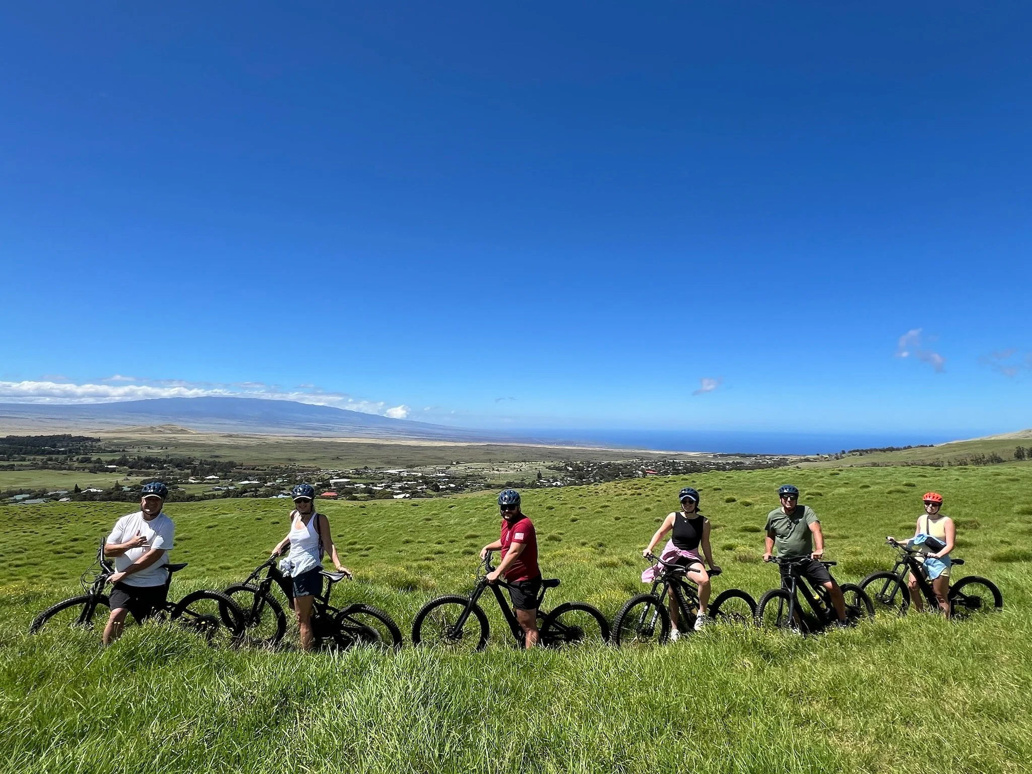 A group of seven people with bicycles standing on a grassy hill under a clear blue sky, with distant mountains and a small town in the background.