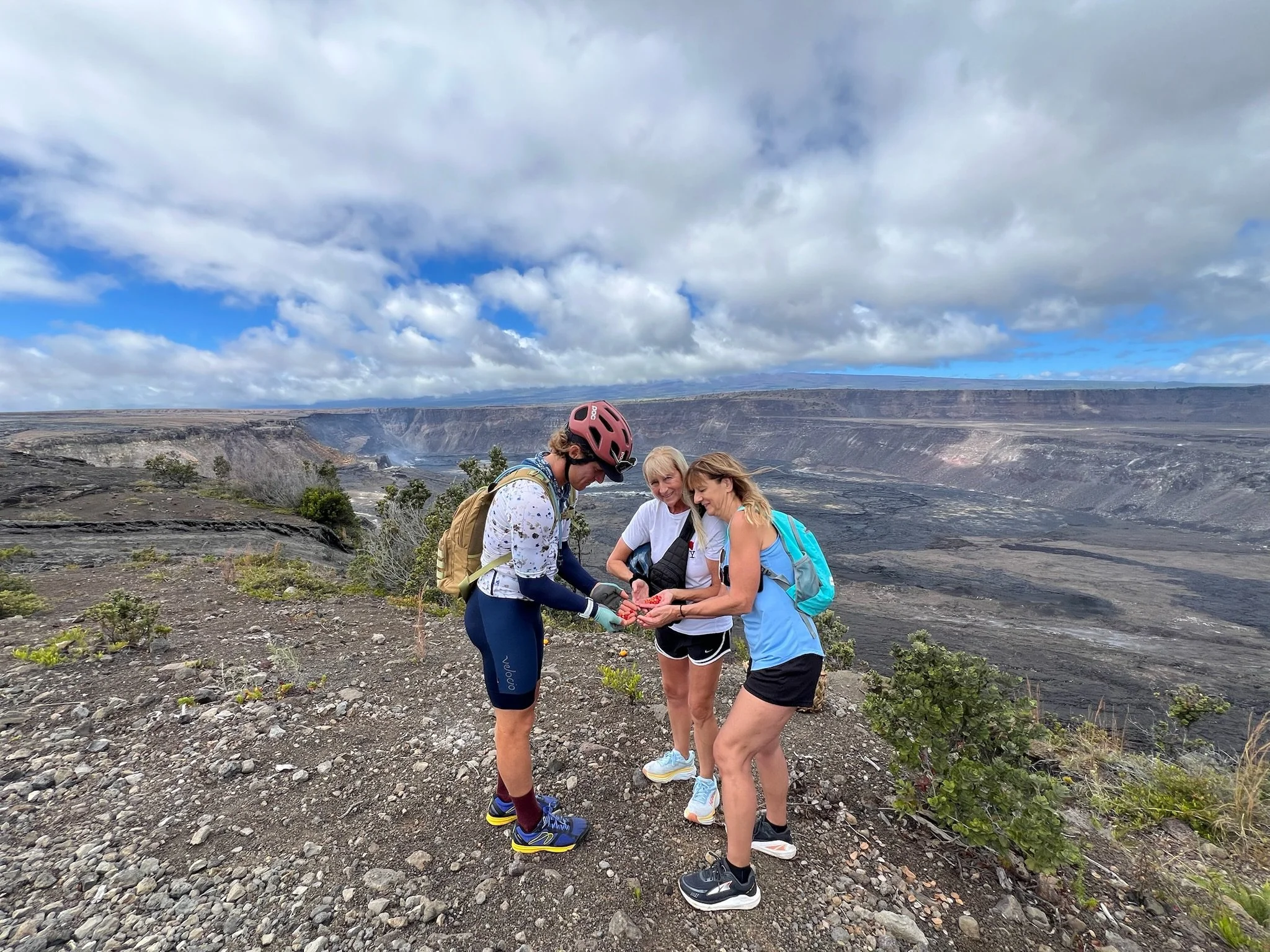 Four women are standing outdoors near a volcanic crater, looking at their phones with a cloudy sky overhead.