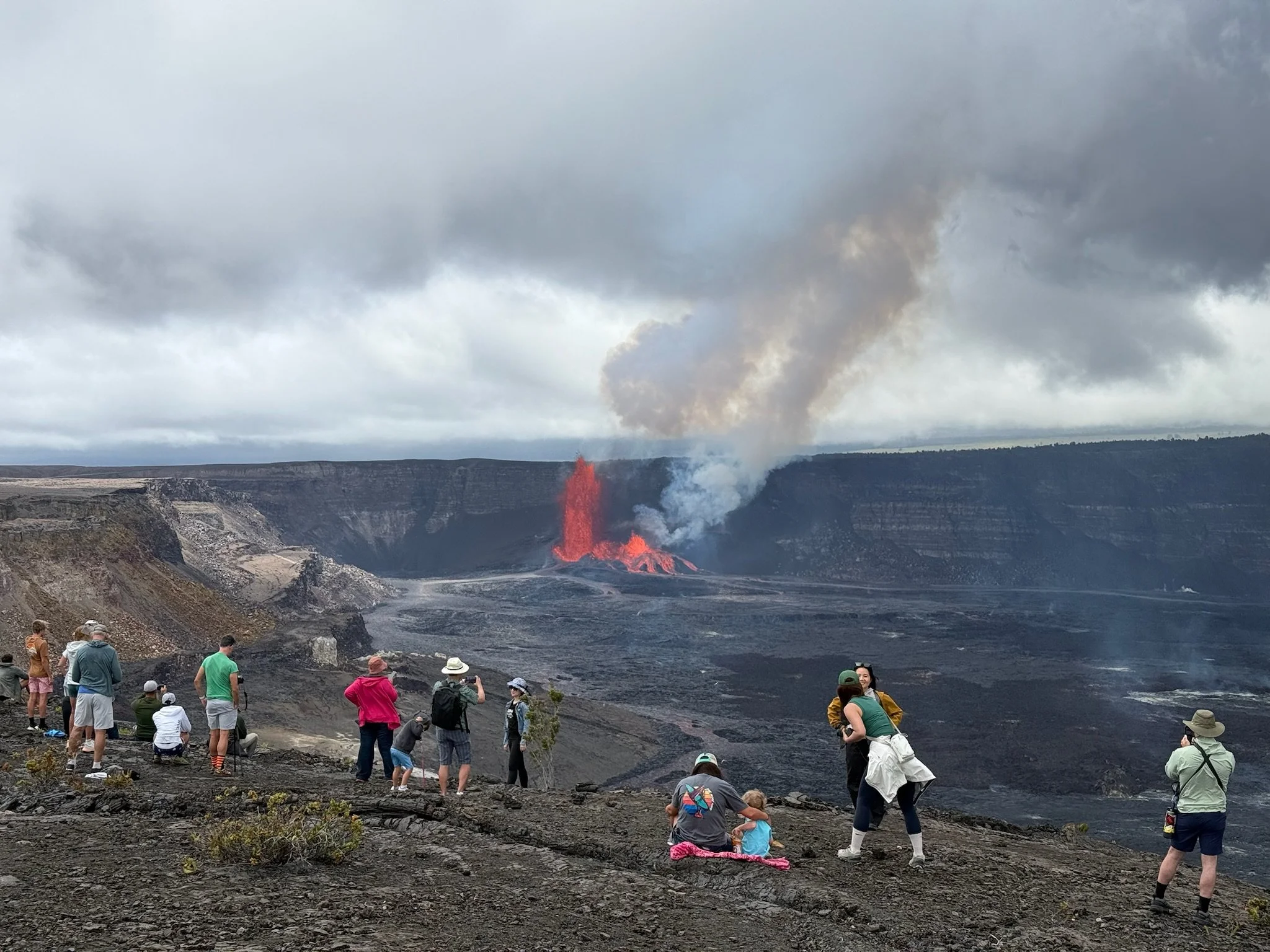 A group of tourists in a volcanic crater watching lava erupt and smoke billowing from the ground under a cloudy sky.