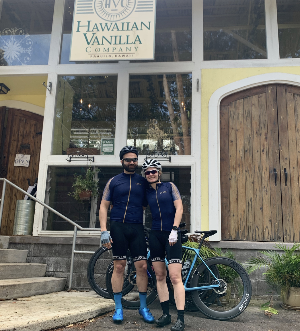A smiling man and woman in cycling gear standing in front of a storefront with wooden doors and large windows. They are wearing helmets, sunglasses, and gloves, with a blue road bike beside them. The store sign above reads 'Hawaiian Vanilla Company' in Paia, Hawaii.