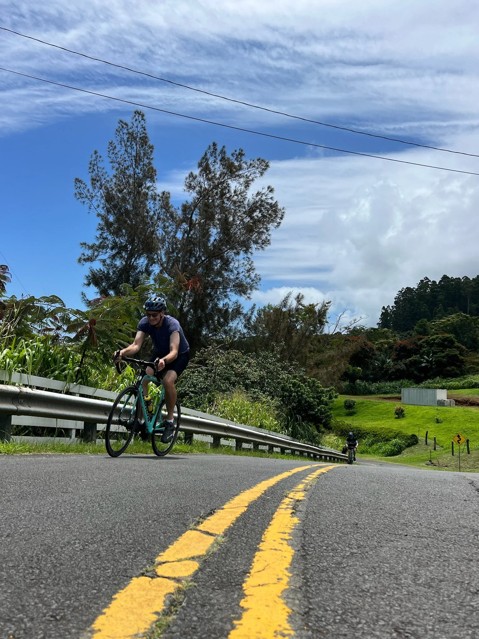Cyclists riding on a winding mountain road with yellow lane markings, greenery, trees, and cloudy sky in the background.
