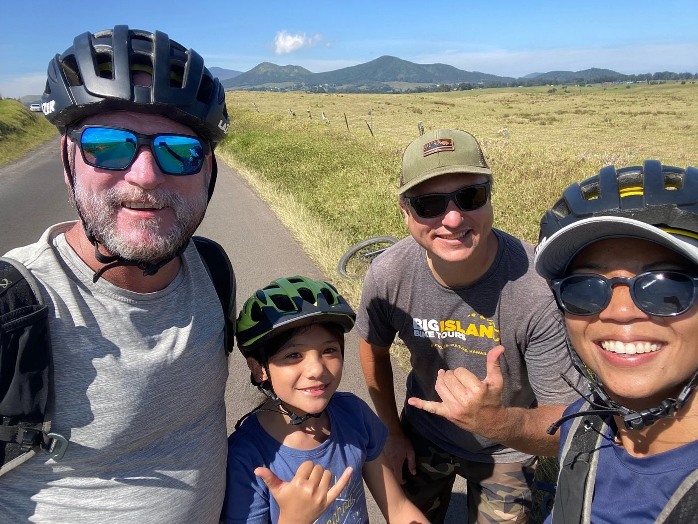 Four people wearing helmets and sunglasses are taking a selfie on a rural road with fields and mountains in the background.