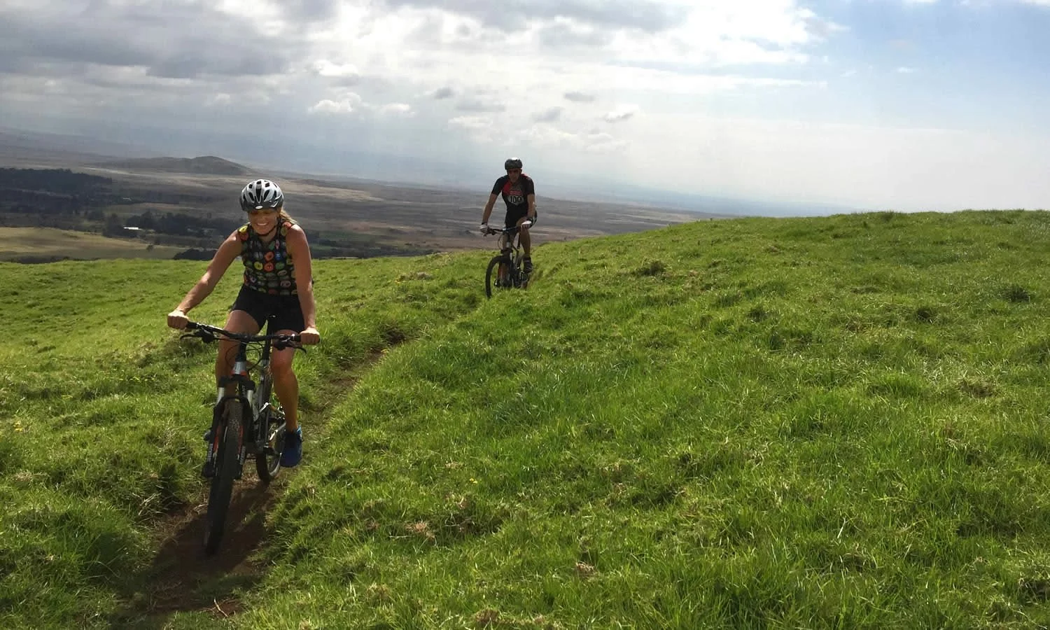 Two people riding mountain bikes on a grassy trail on a hill, with a scenic landscape and cloudy sky in the background.