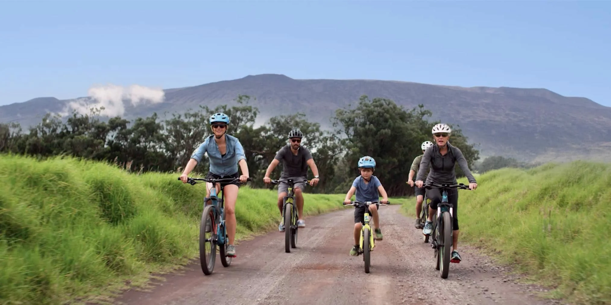 A group of people, including children and adults, riding mountain bikes on a dirt trail through green fields with trees and a mountain in the background.