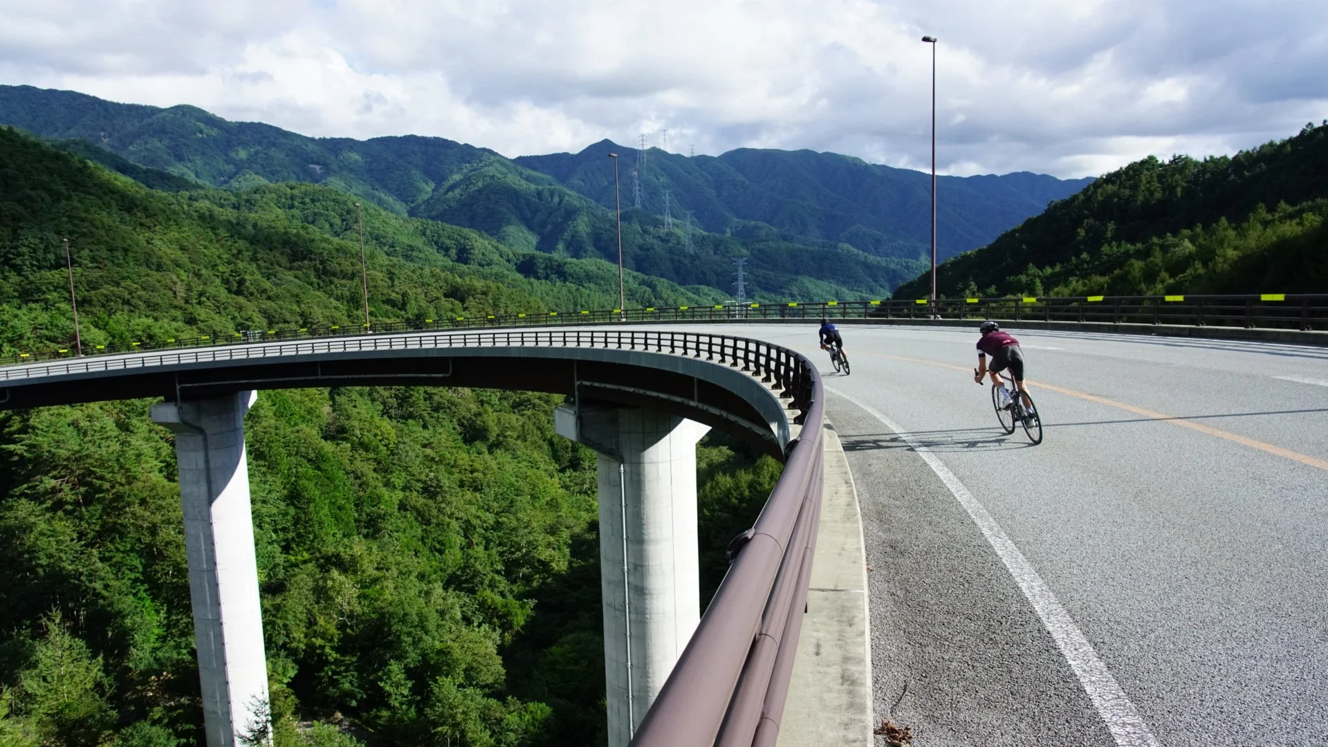 Two cyclists riding on a curved mountain road with green hills and cloudy sky in the background.