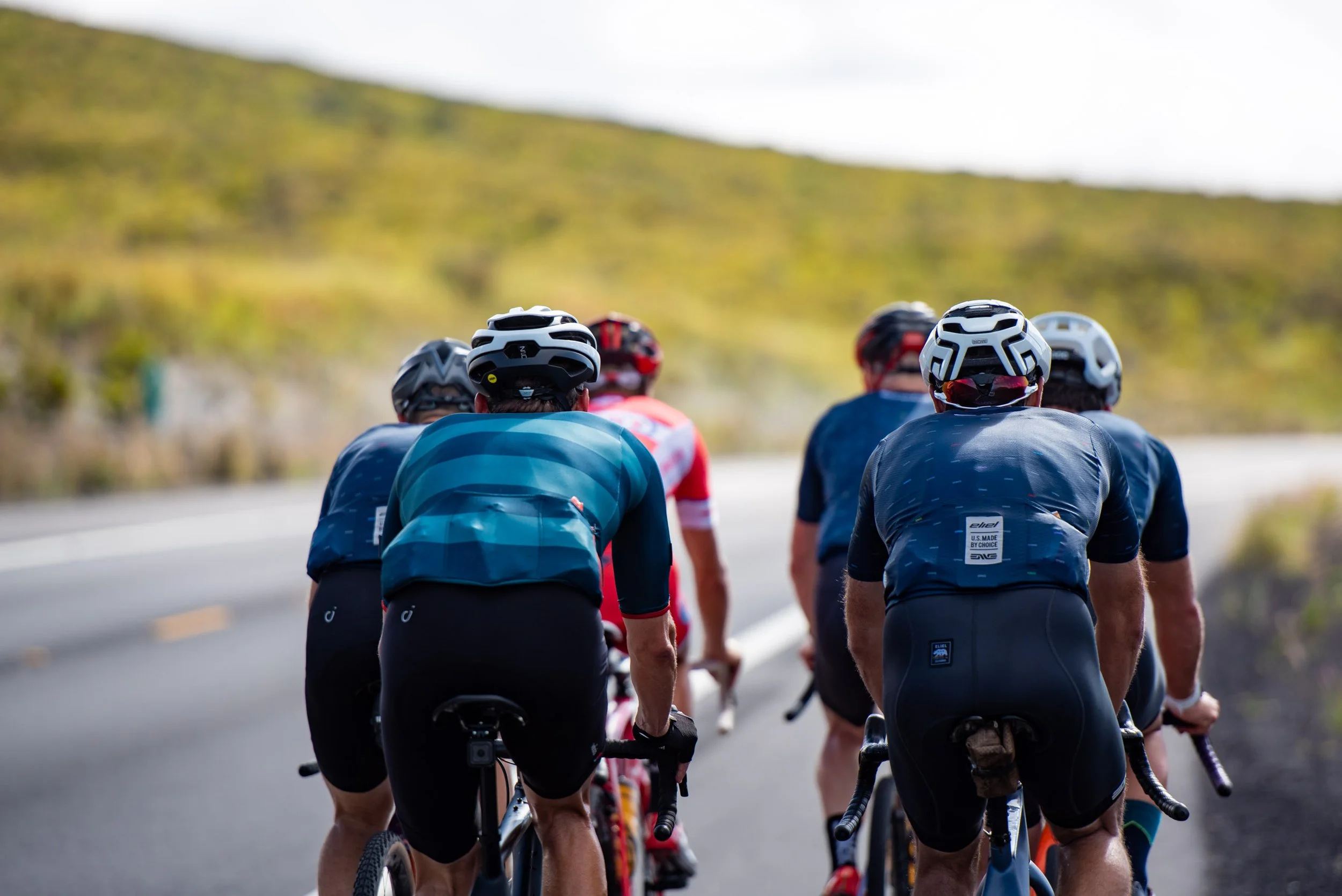 A group of cyclists riding on a road in a rural, hilly area, seen from behind, wearing helmets and athletic clothing.