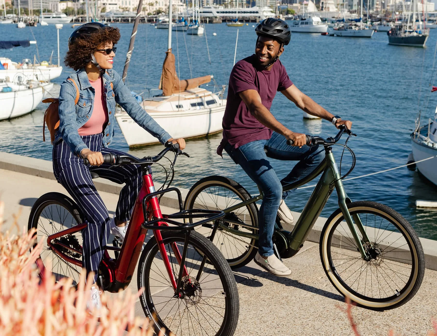 Two people riding electric bikes along a marina with sailboats docked in the background on a sunny day.