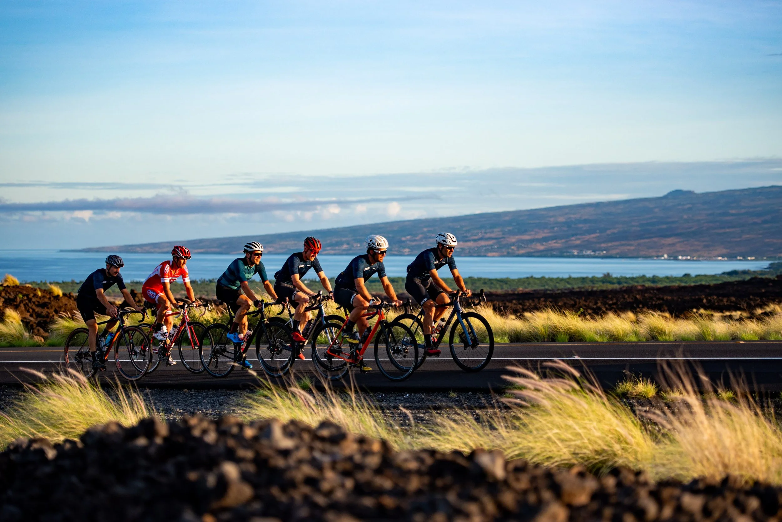 A group of cyclists riding on a road beside a body of water with hills in the background under a clear sky.