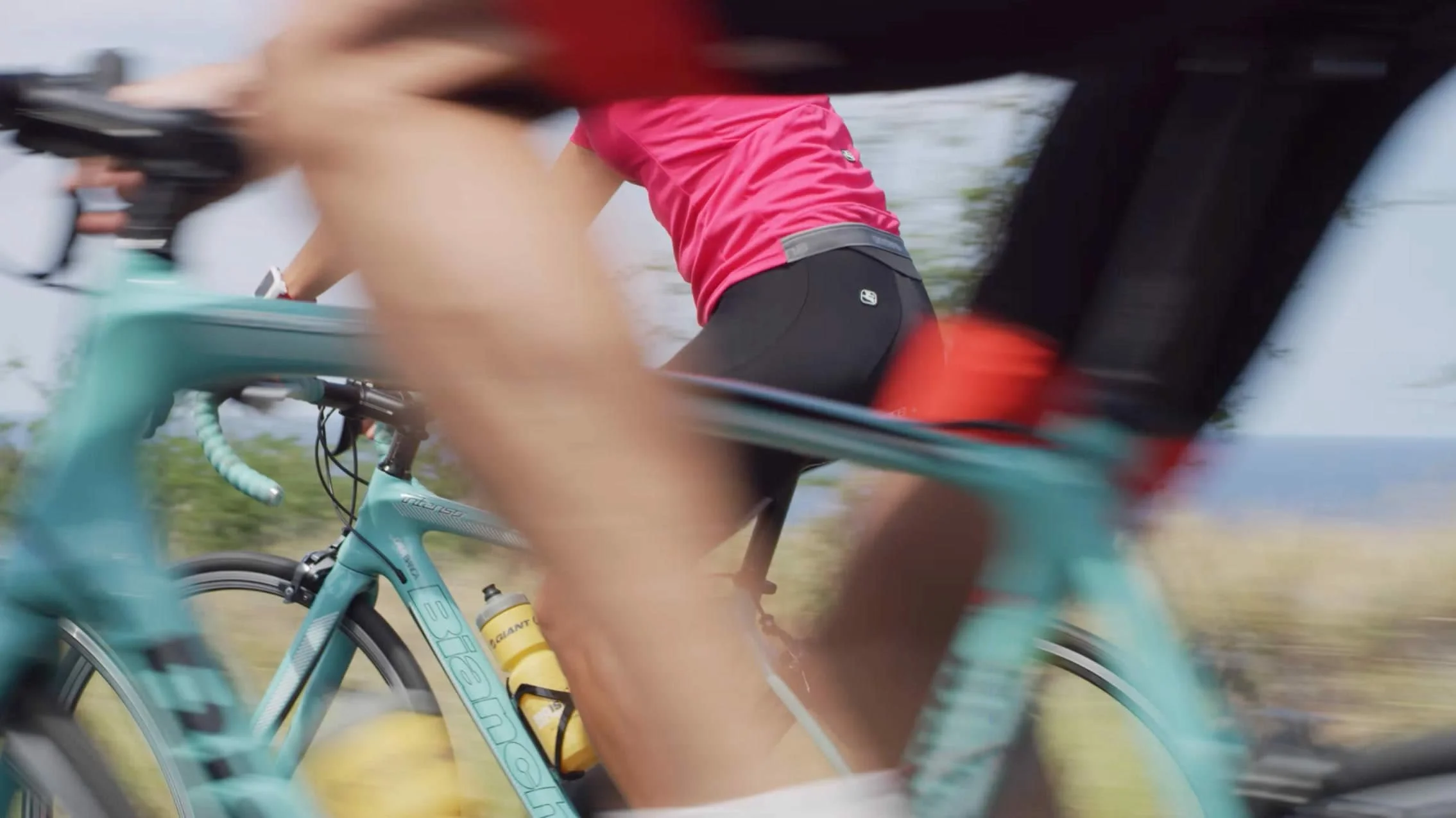 Close-up of cyclists riding on a trail, with one rider wearing a pink shirt and black shorts, and a turquoise bike visible in the foreground.