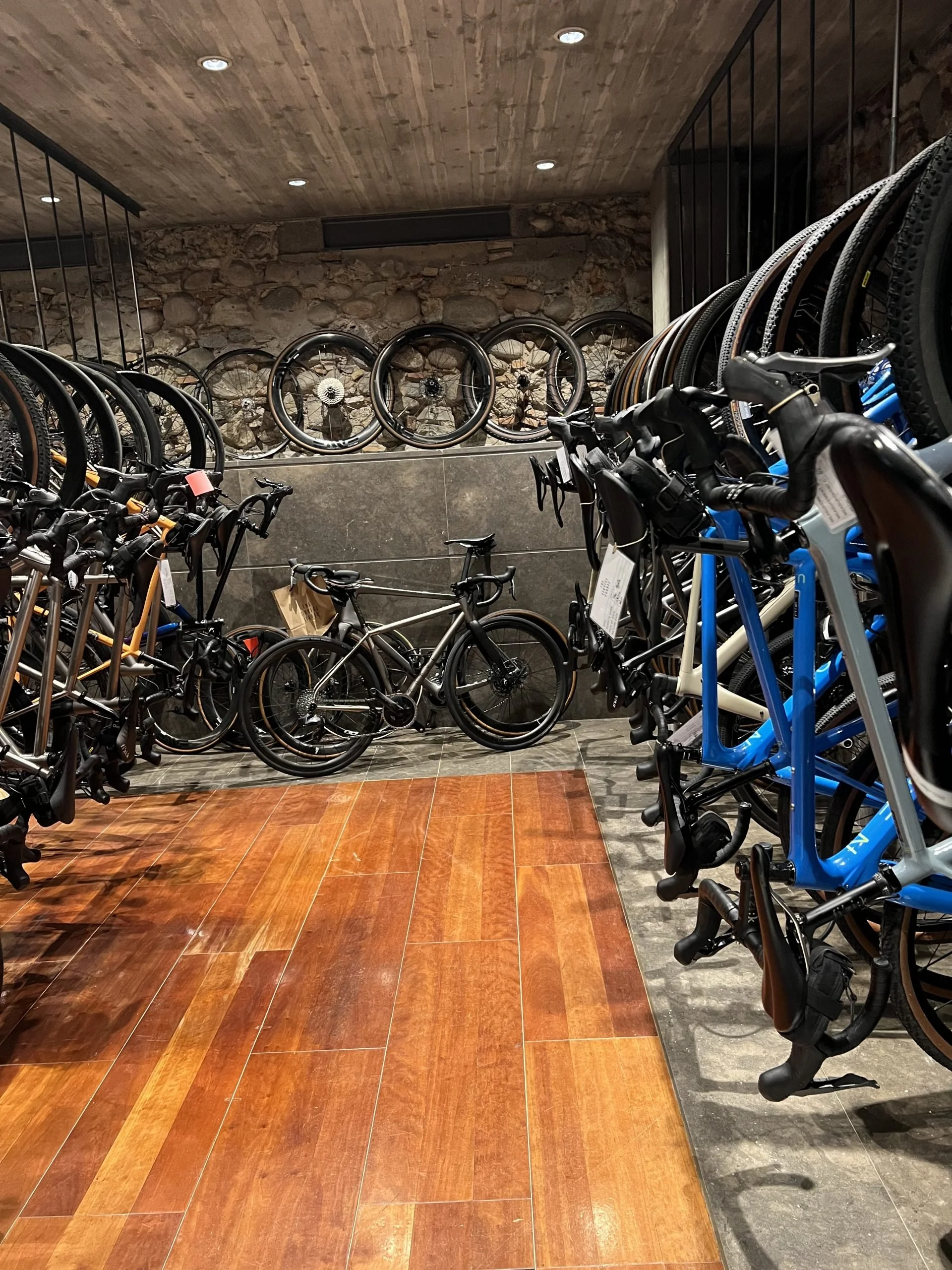 Indoor bike shop with rows of bicycles on display, wood and stone interior, ceiling lights, and a stone wall in the background.