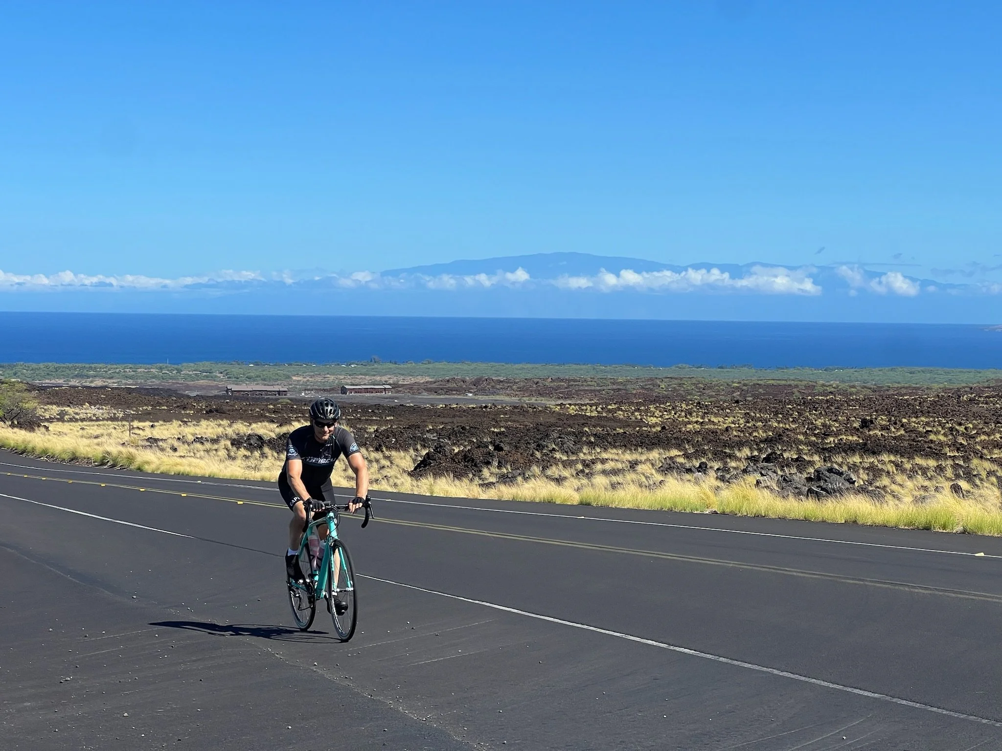 A cyclist riding on a paved road with a scenic landscape of ocean, mountains, and sky in the background.