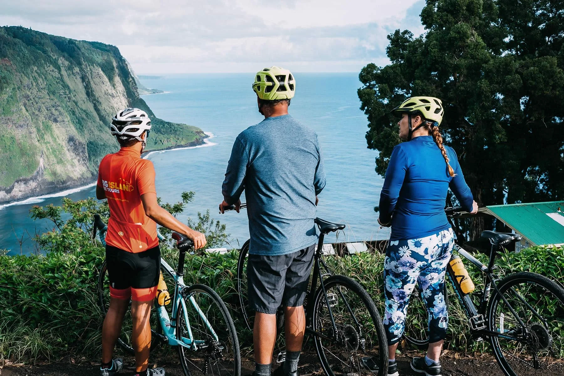 Three cyclists, two women and one man, standing with their bikes overlooking a coastal landscape with cliffs, ocean, and trees, all wearing helmets.
