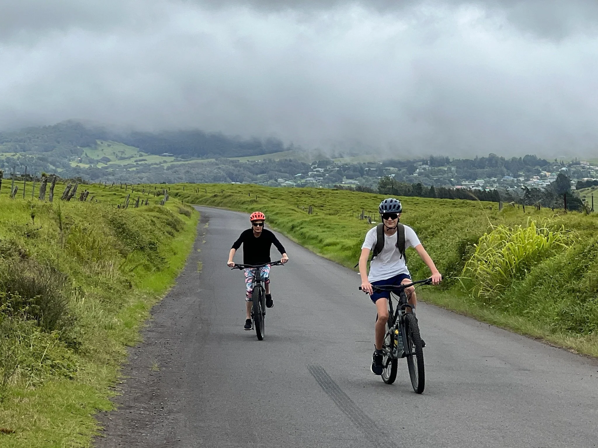 Two children riding bicycles on a countryside paved path under cloudy skies, with green fields and a small town in the background.