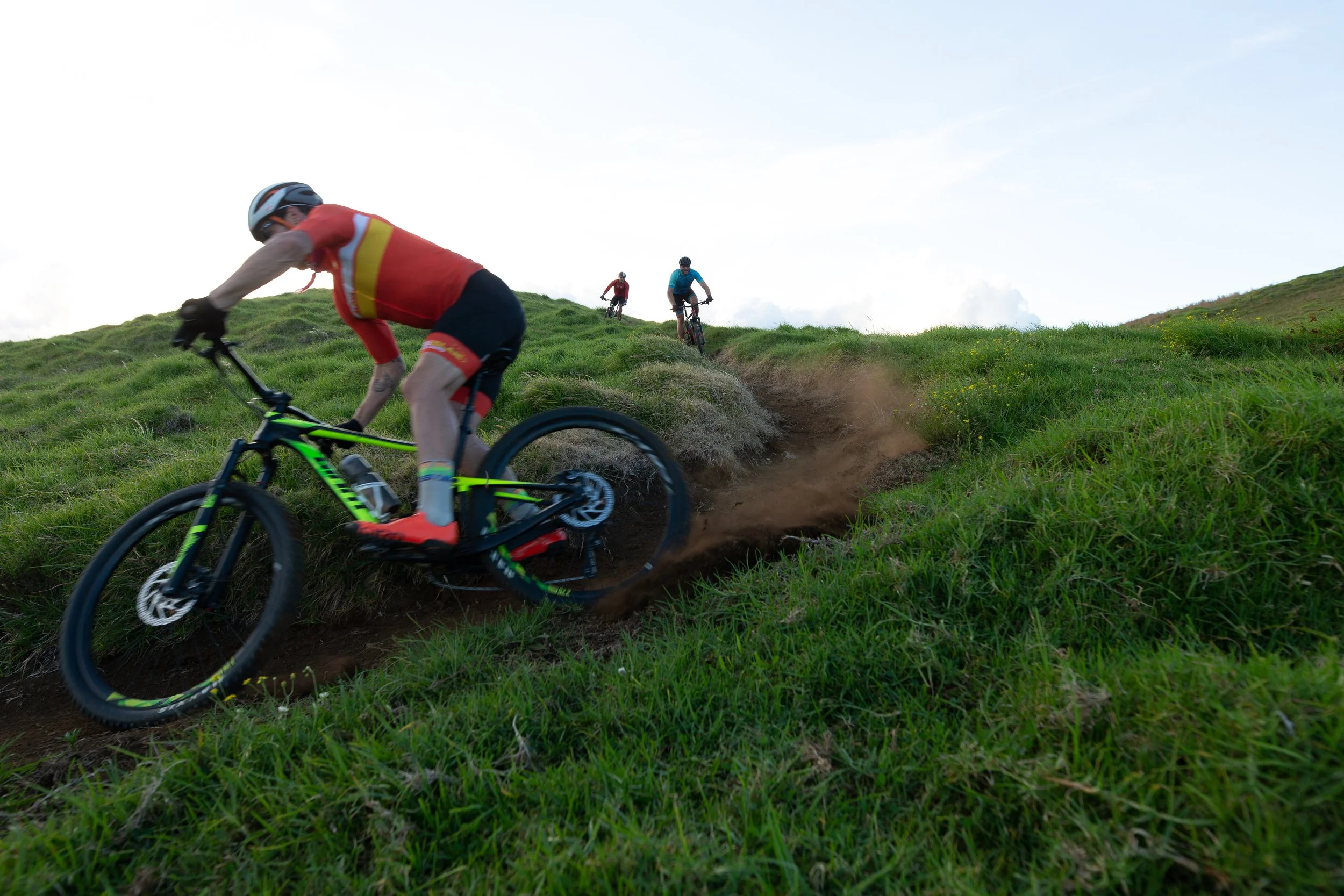 Three mountain bikers riding on a trail through lush green rolling hills with a cloudy sky overhead.