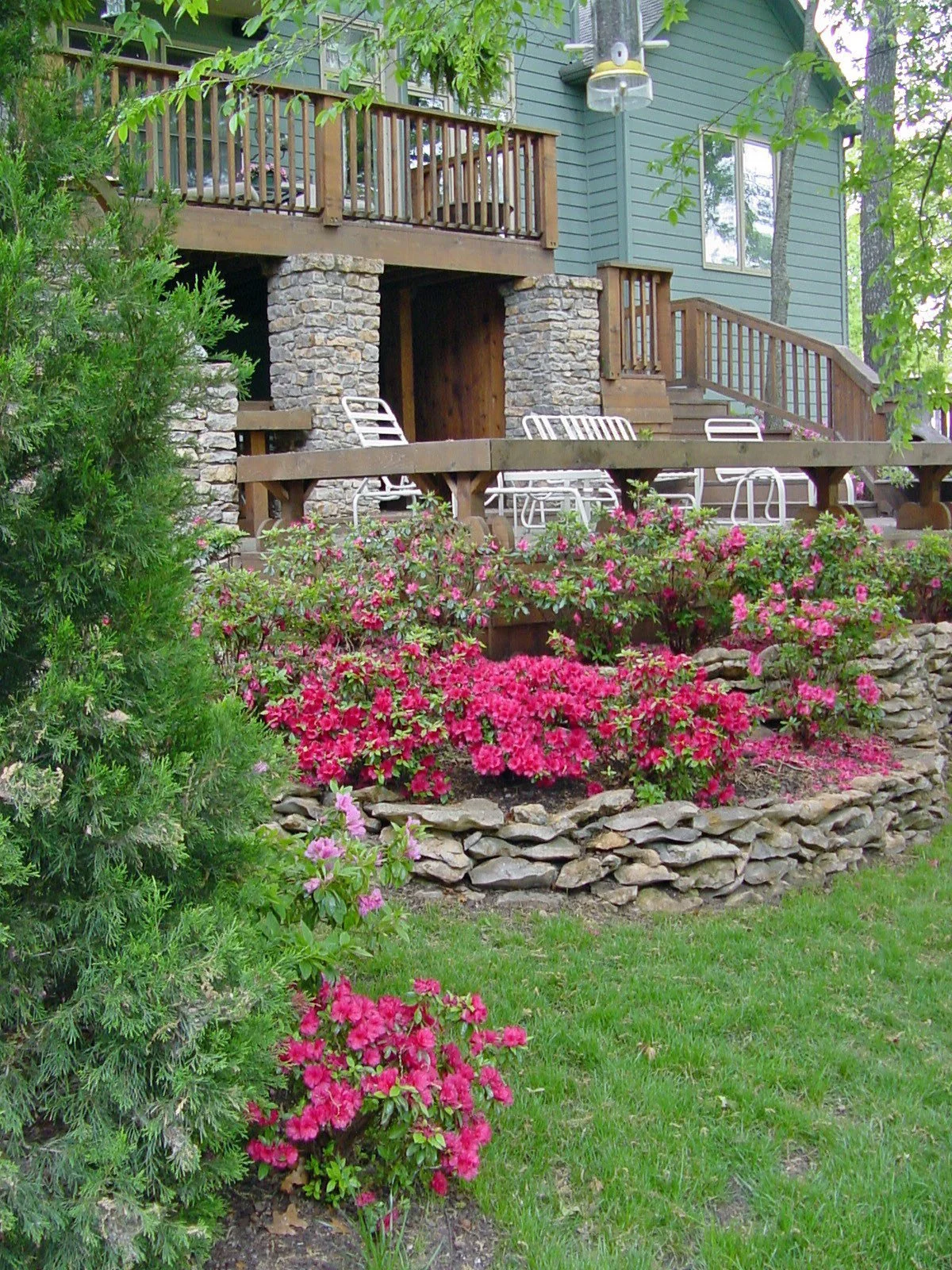 A house with a wooden porch and stone pillars, surrounded by green trees and pink flowers in a garden.