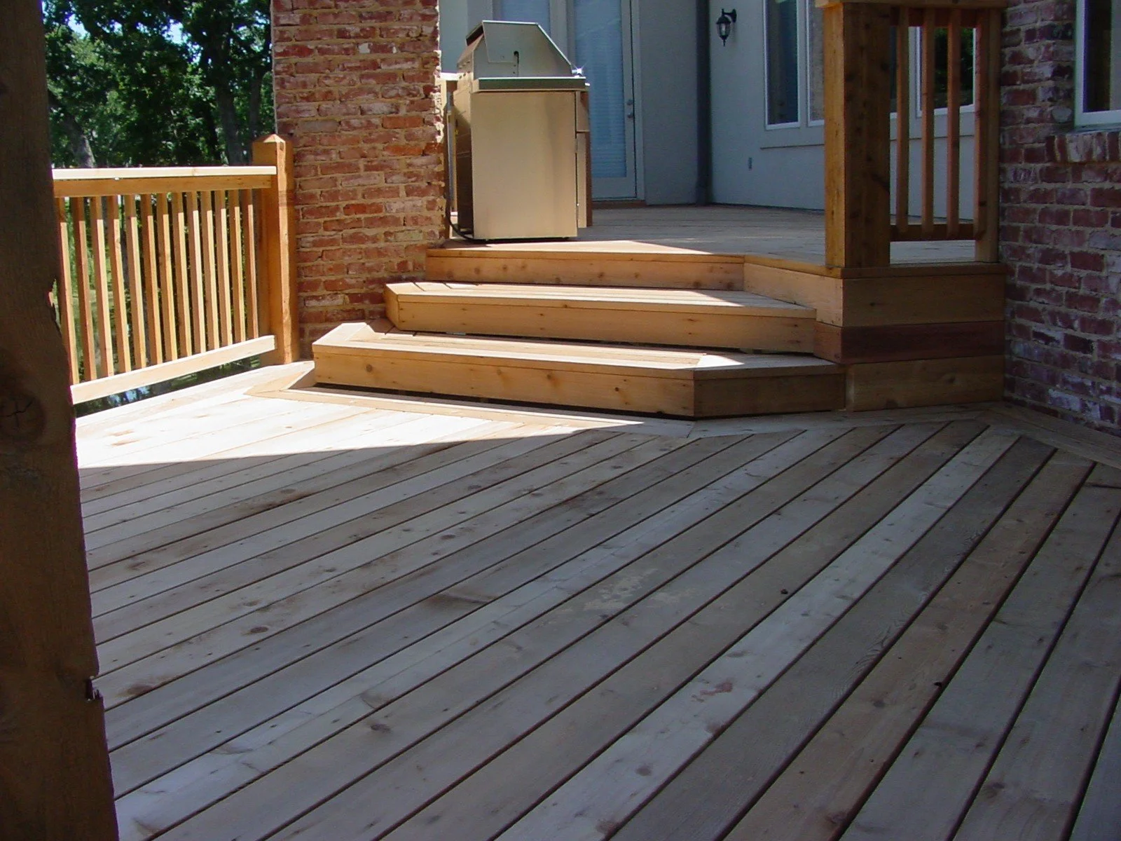 A newly built wooden deck with stairs leading up to a screened porch on a house, outdoor grill, brick wall, wooden railing, and trees in the background.