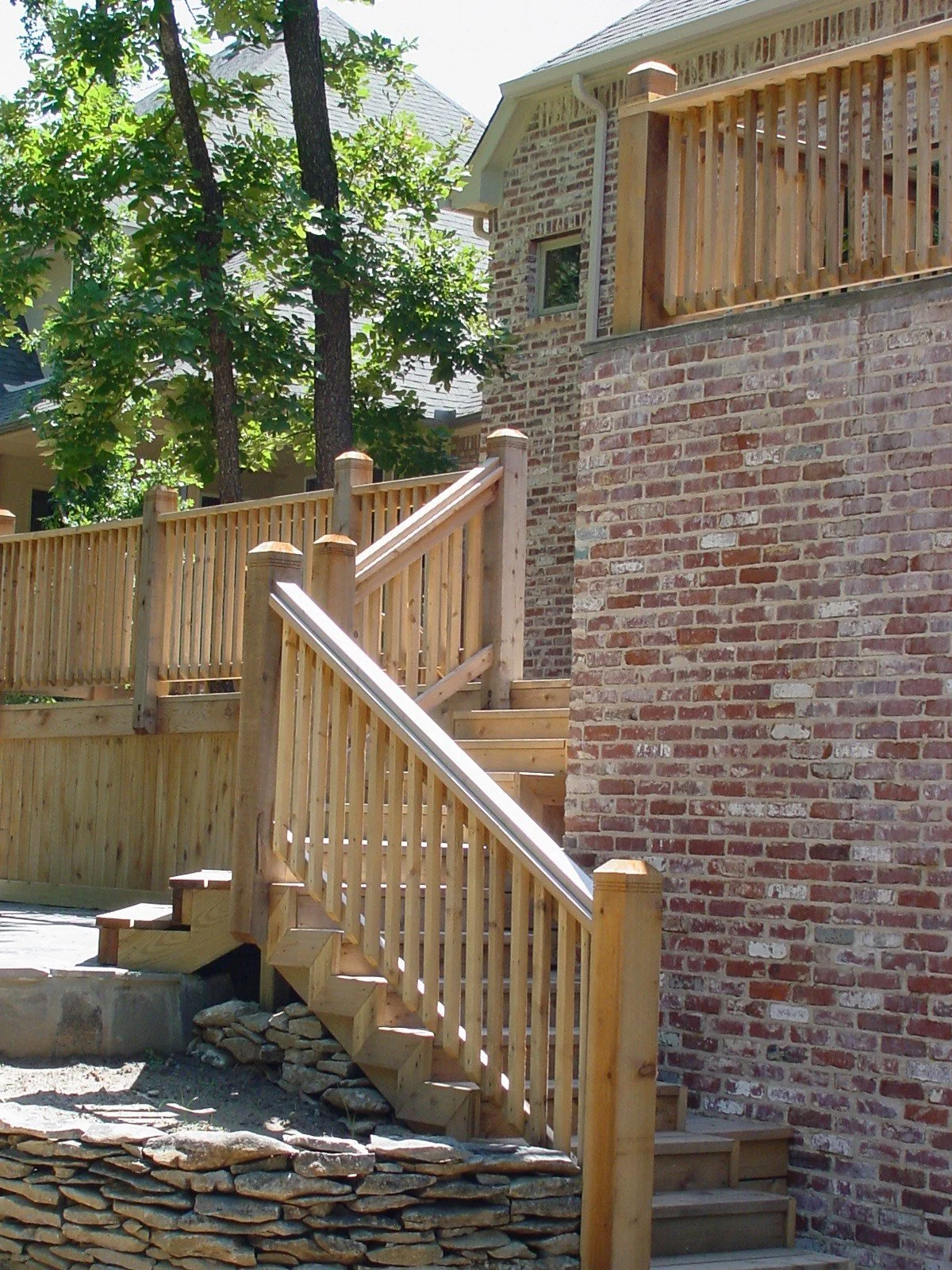 Newly built wooden staircase with railing attached to a brick house, surrounded by green trees.