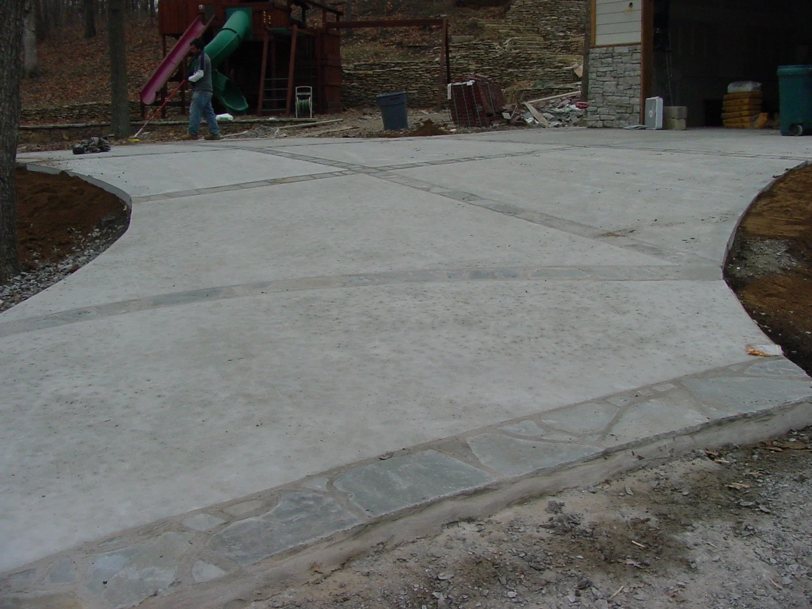 A concrete sidewalk in front of a house, with a child playing with a garden hose, and a play structure with slides in the background.
