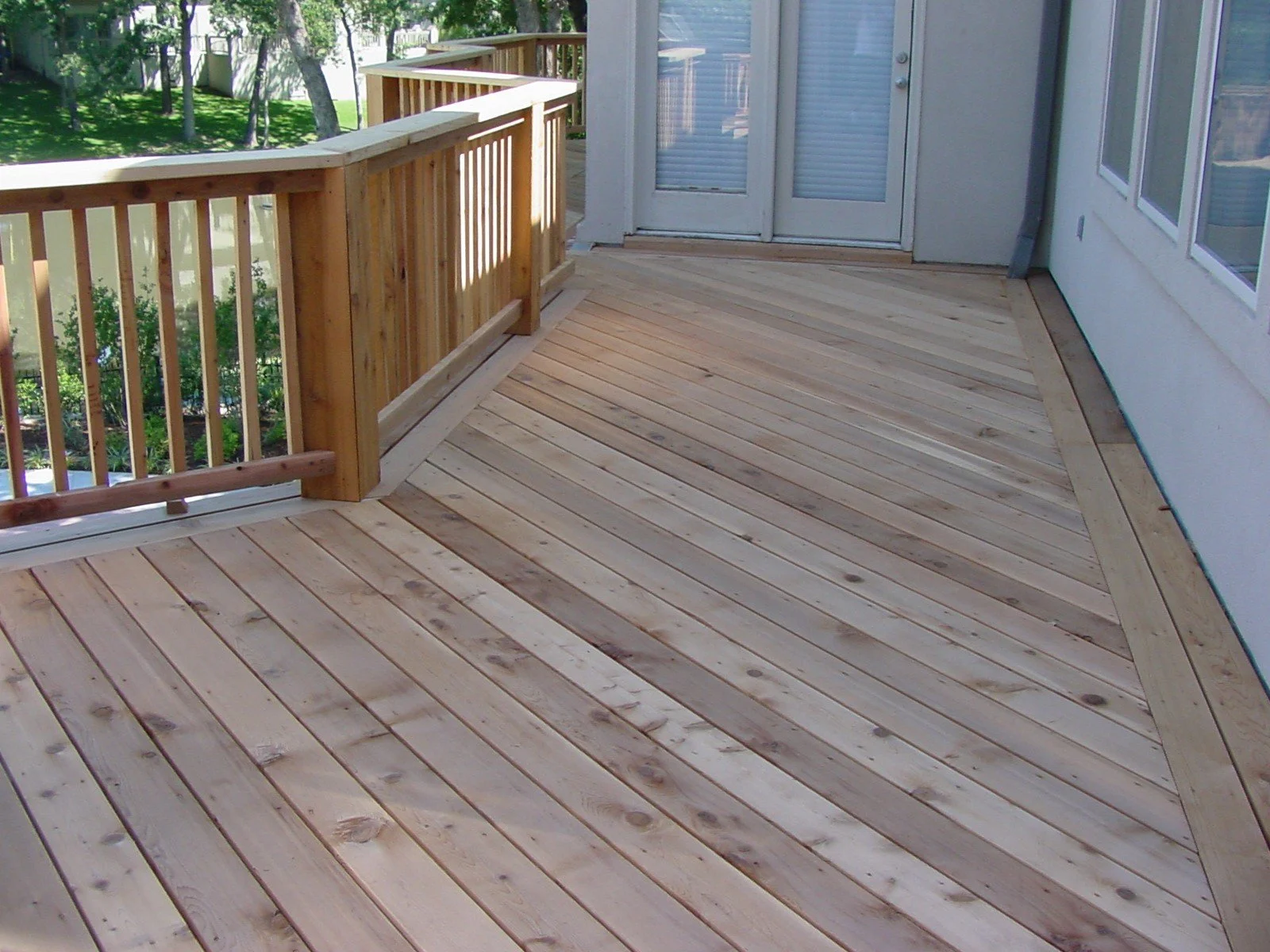 A wooden deck with a railing and a set of white sliding glass doors on a house exterior. The deck is surrounded by a backyard with green grass and trees.