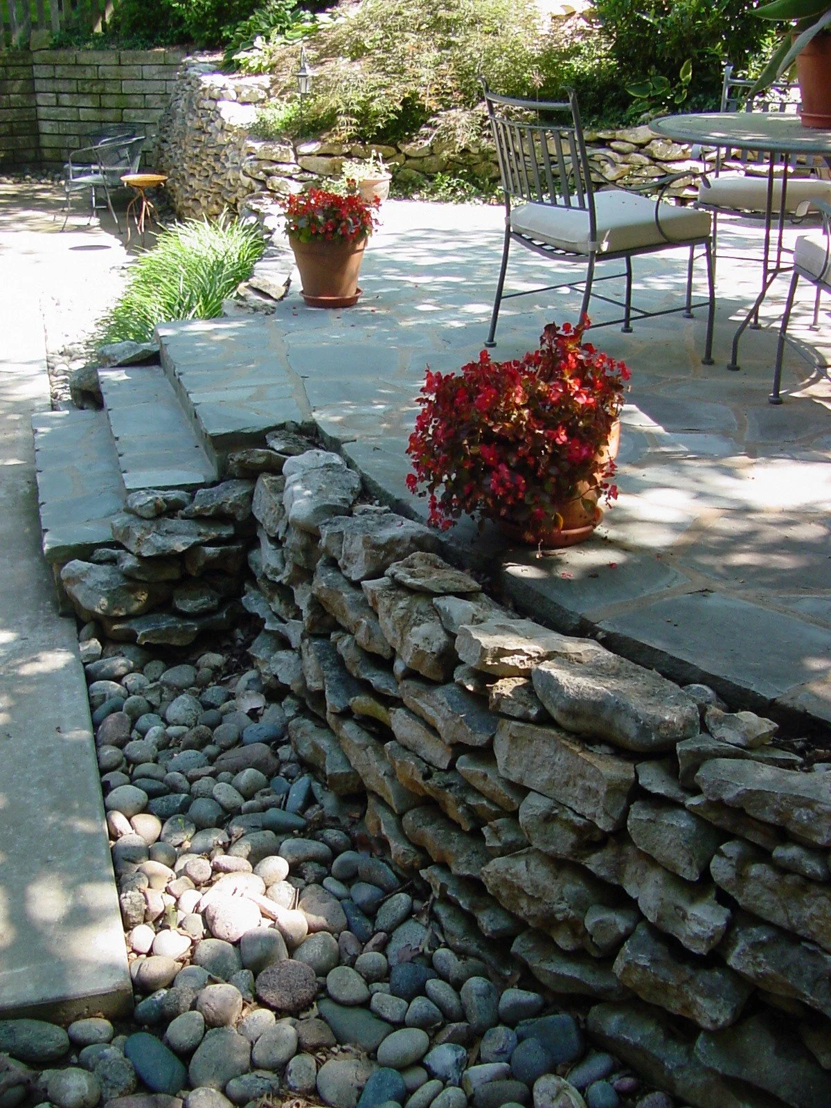 A backyard patio with a stone retaining wall, potted plants with red flowers, a table and chairs, surrounded by greenery and shaded by trees.