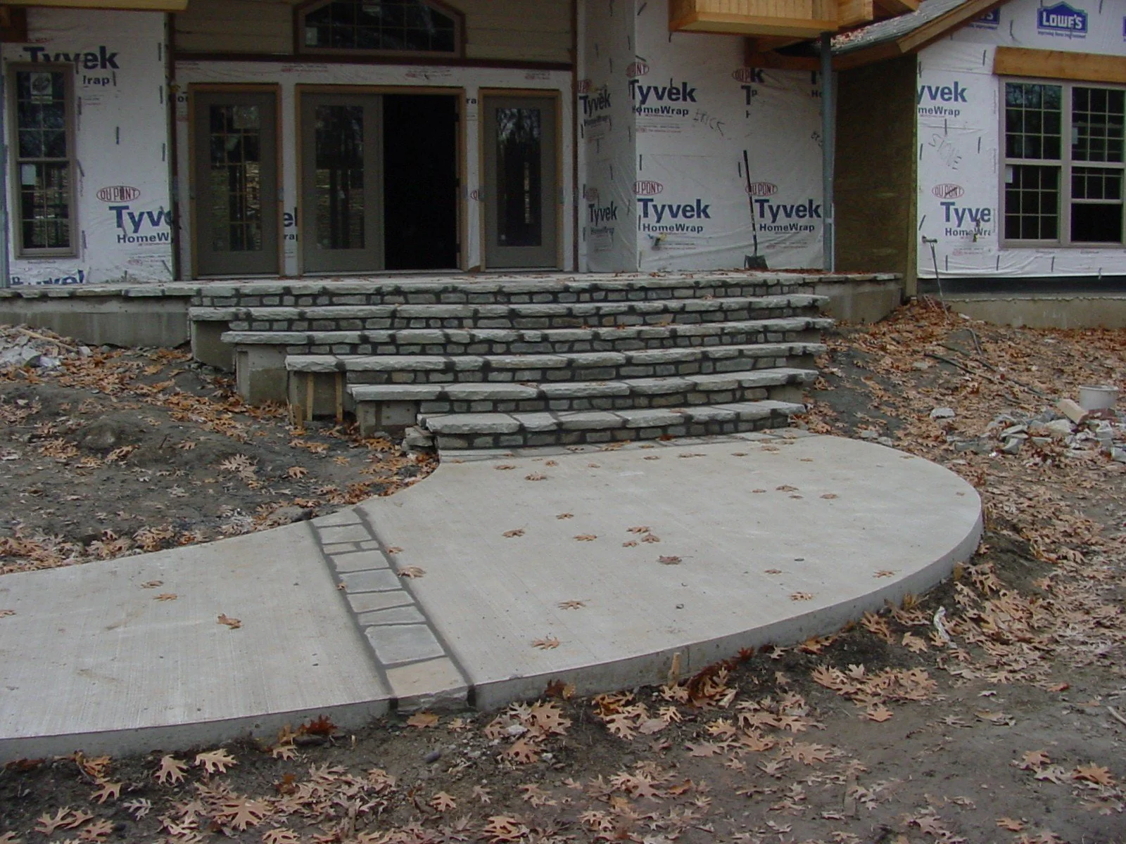Construction site of a house with a front porch and stone steps, surrounded by dirt and fallen leaves, with partially installed windows and exterior wall sheathing.