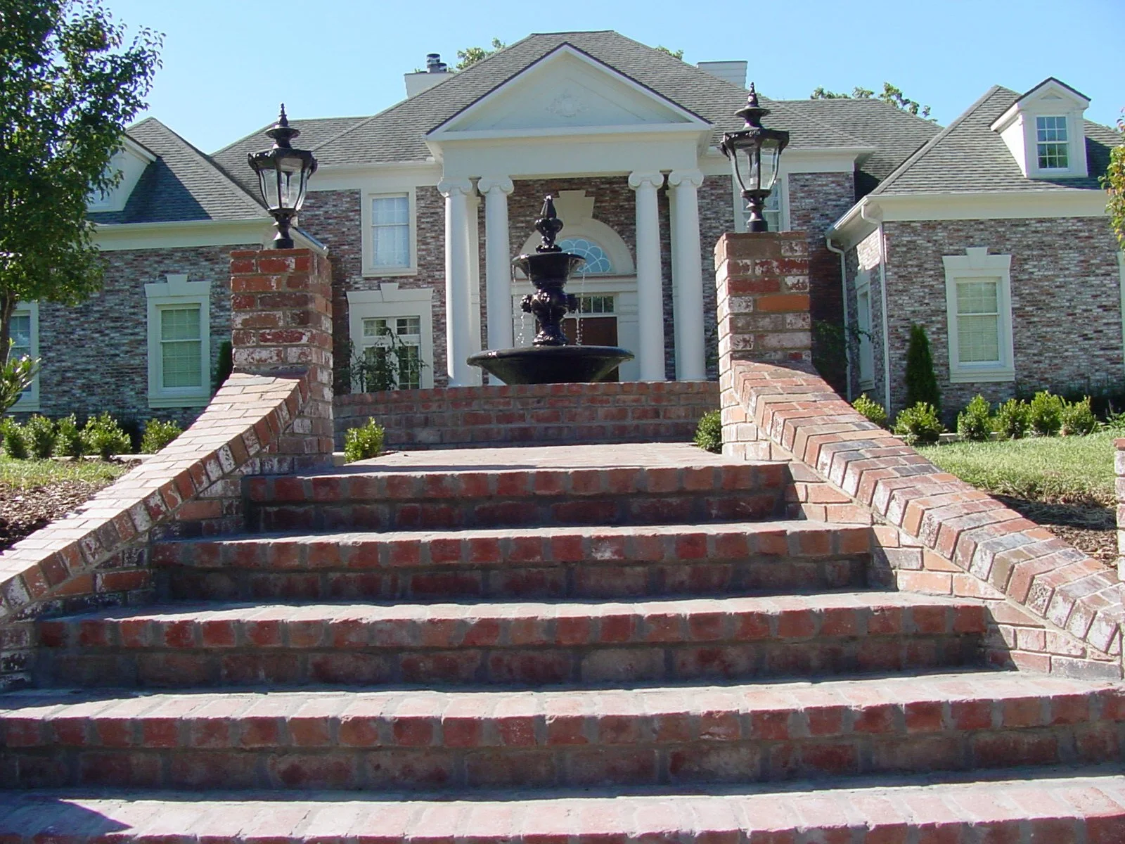 A brick staircase leading up to a large house with a fountain and black lampposts in front, surrounded by garden landscaping.