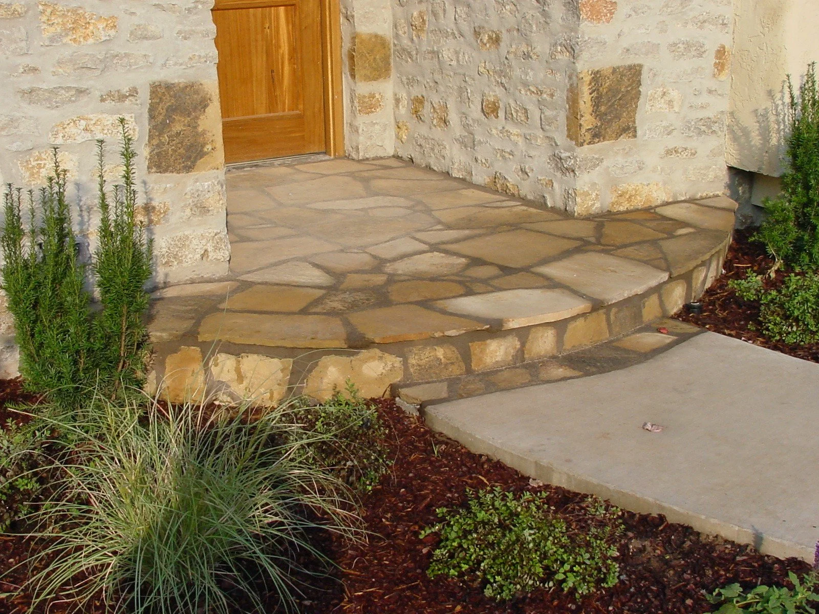 Front porch of a house with a stone and brick step landing, wooden door, surrounded by bushes and mulch.