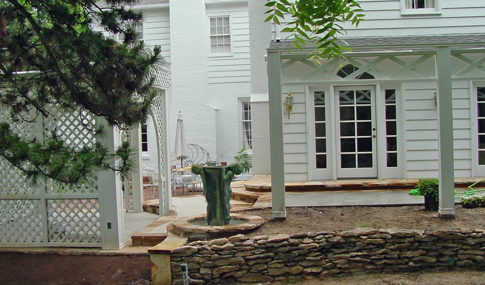 Backyard with white house, patio area with chairs and umbrella, garden fountain, stone steps, and a garden bed bordered by stacked stones.