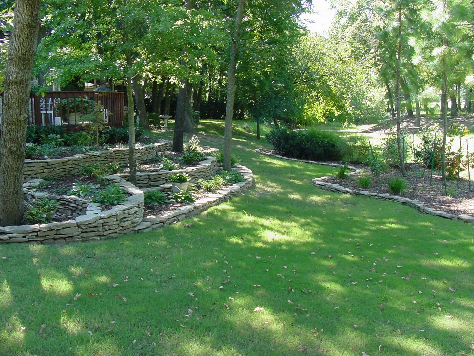 A well-maintained backyard garden with tiered stone garden beds, green grass, and lush trees providing shade.