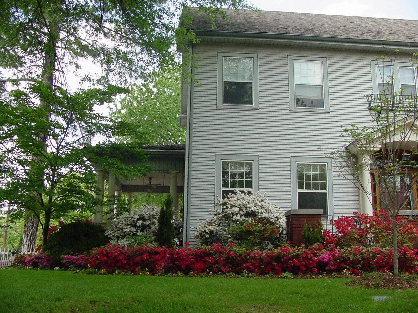View of a multi-story house with grey siding, multiple windows, a porch with columns, and a garden with colorful flowers and green trees.