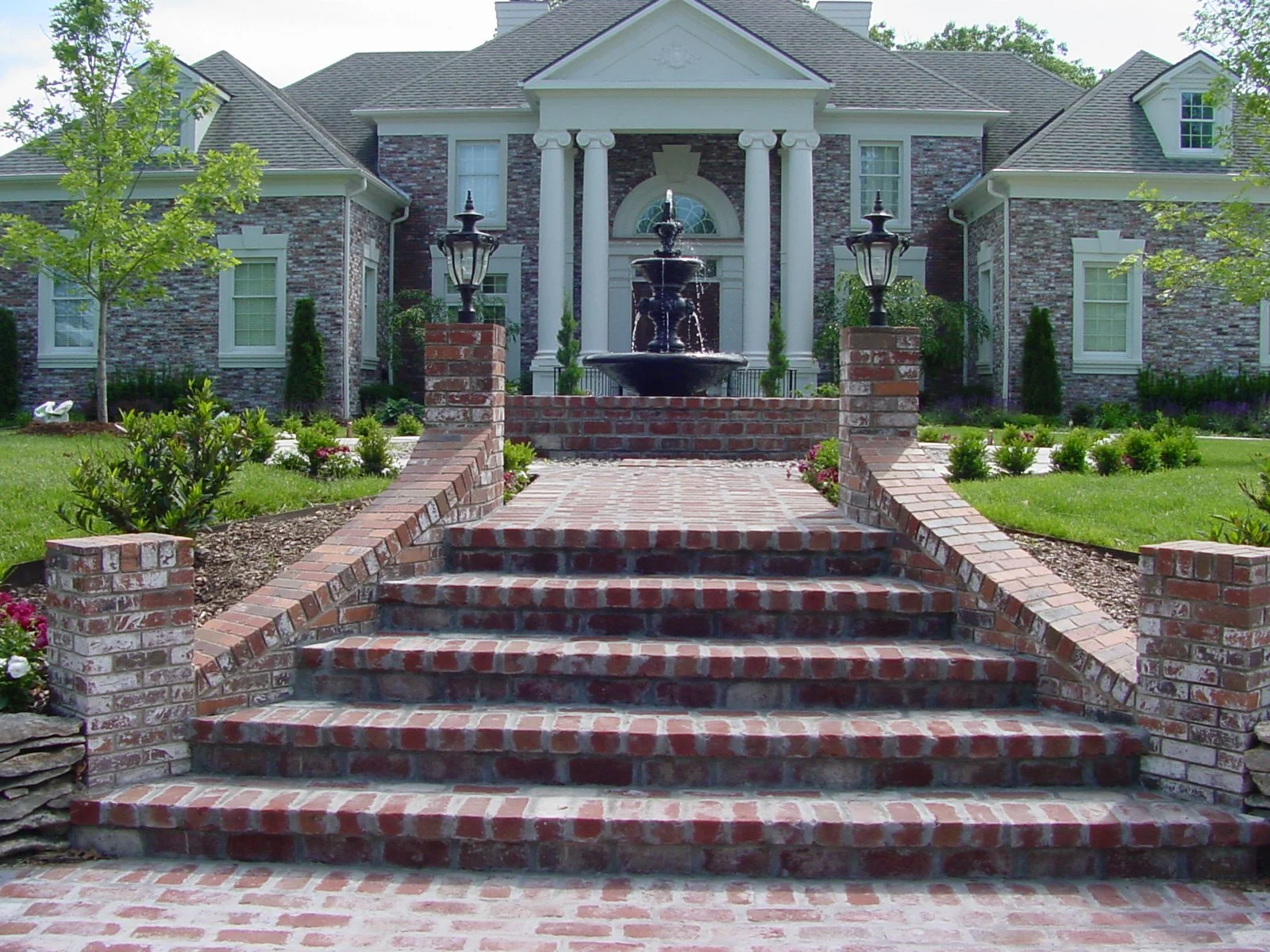 Brick stairs leading to the entrance of a large, elegant house with a fountain and garden in front.