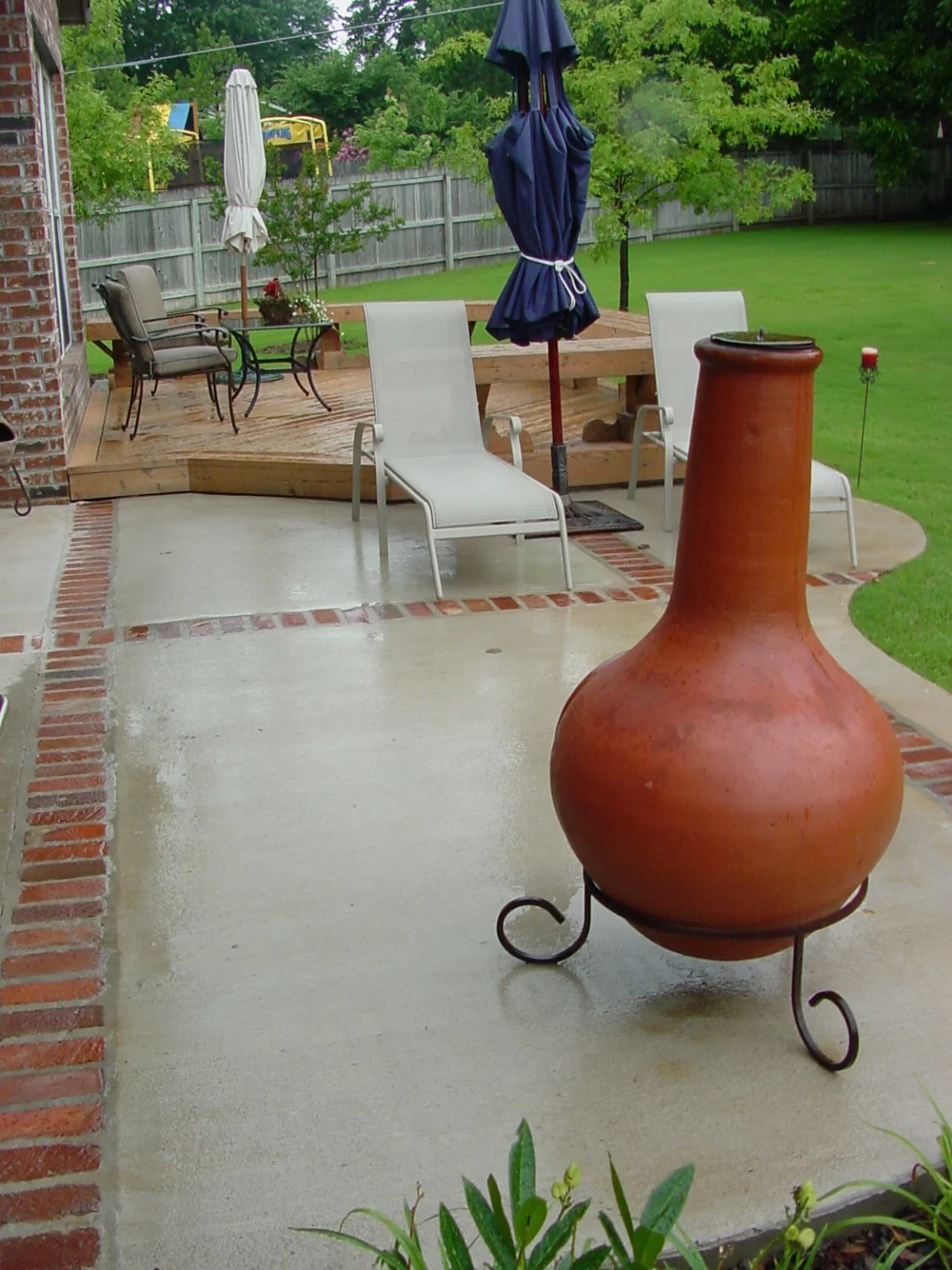 Back patio area with brick and concrete flooring, outdoor chairs, white and blue patio umbrellas, a large terracotta chiminea on metal stand, and a green yard with trees and a wooden fence in the background.