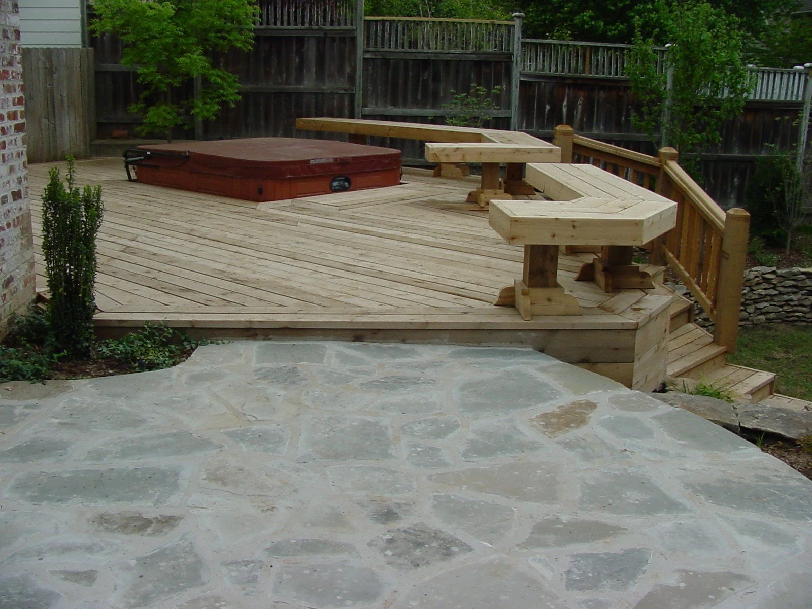 A backyard wooden deck with steps leading up to it, surrounded by a stone patio and a wooden fence. There is a hot tub on the deck and some wooden benches.