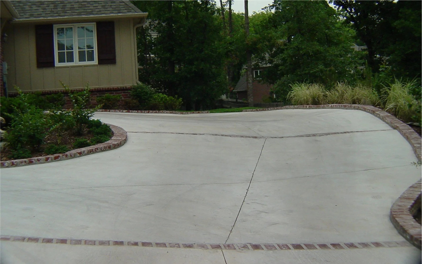 A curved residential driveway with a brick border, surrounded by small plants and bushes, leading to a house with beige siding and a window with shutters, with large trees in the background.