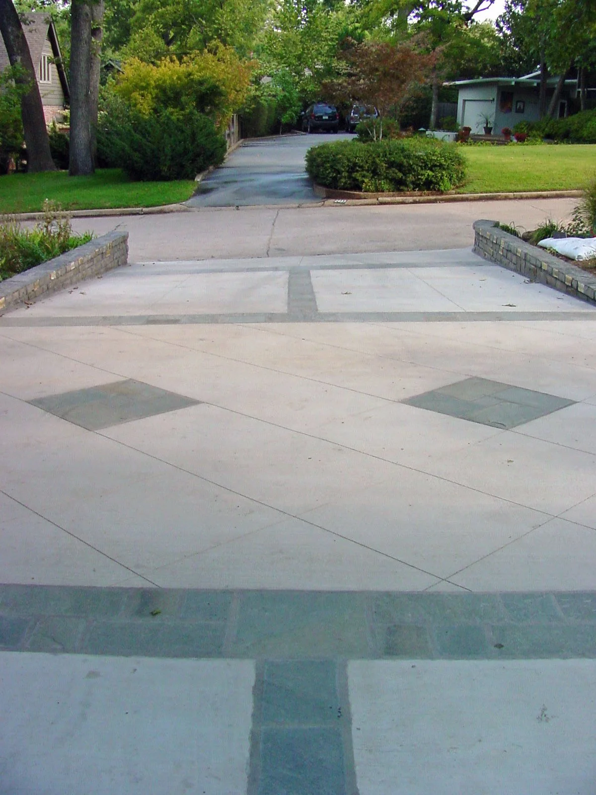 A residential driveway with a brick border and decorative gray tiles leading to a street with parked cars, surrounded by green trees and shrubs.