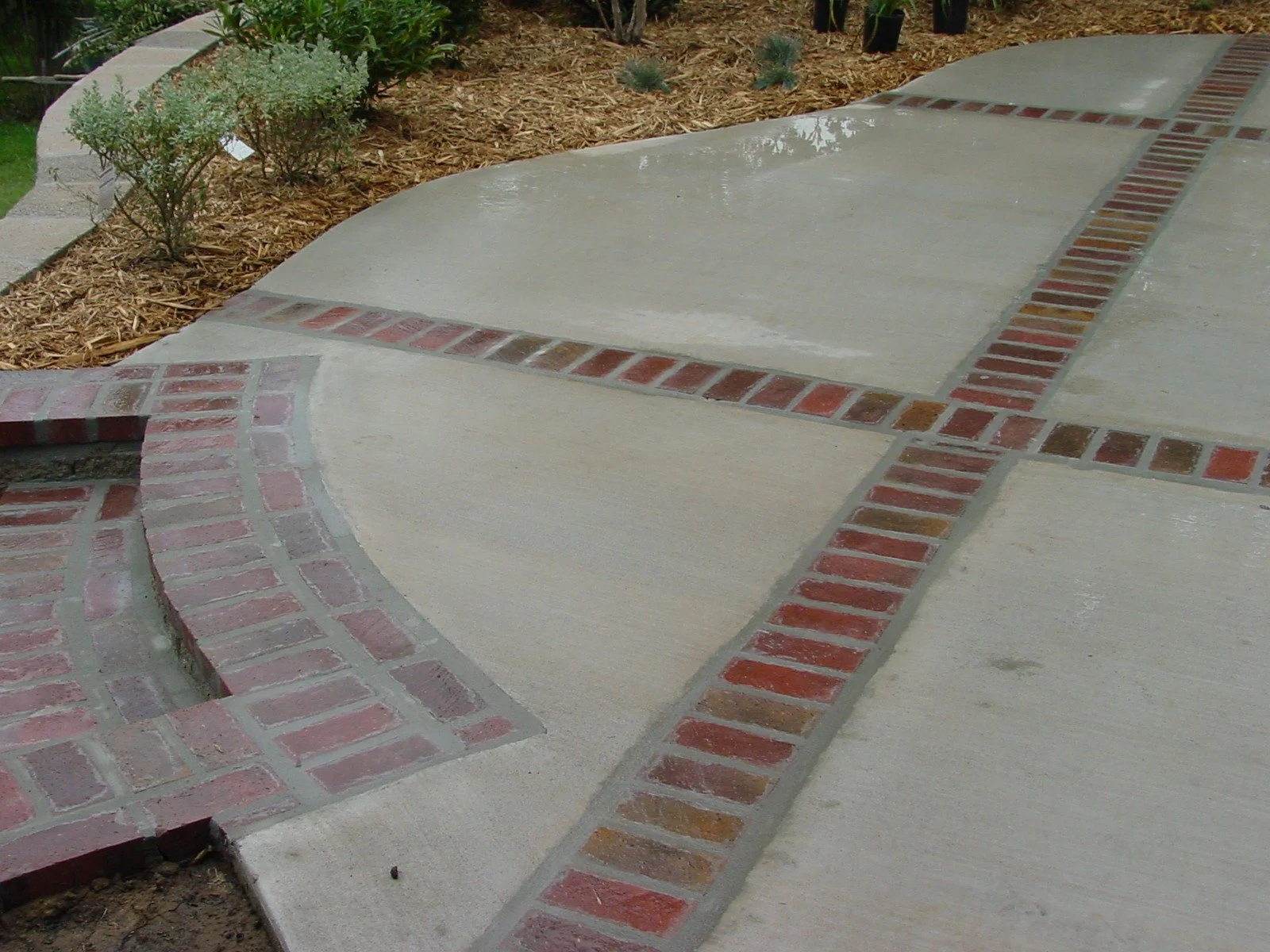 Close-up of a concrete sidewalk with a decorative brick pattern featuring red and brown bricks, adjacent to a garden bed with mulch, small bushes, and potted plants.