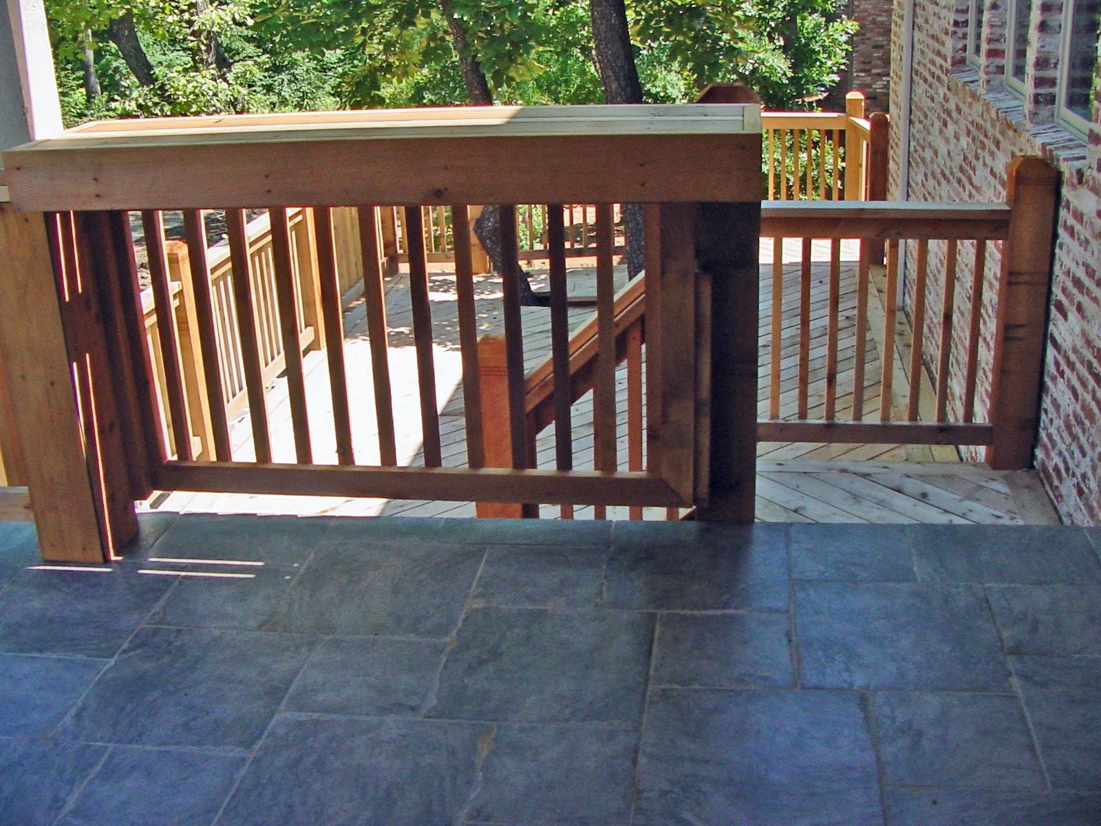 A wooden outdoor balcony with a railing and stairs leading down to a lower level, with trees and foliage in the background and a brick wall on the right side.