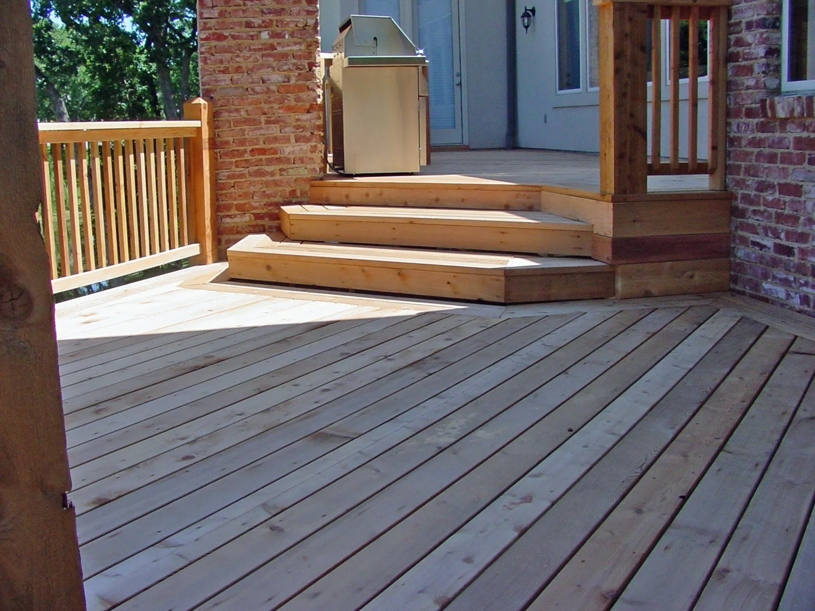 View of a wooden deck with stairs leading up to a patio area, featuring a brick wall on one side and a railing on the other, with trees and a house with a sliding glass door in the background.