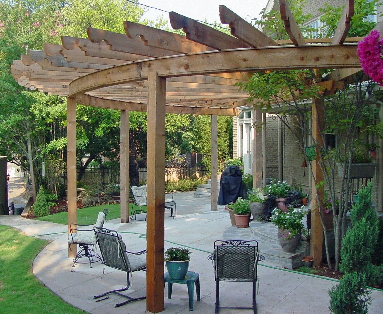 A backyard patio with a wooden pergola, potted plants, patio furniture, and a lawn with trees and shrubbery.