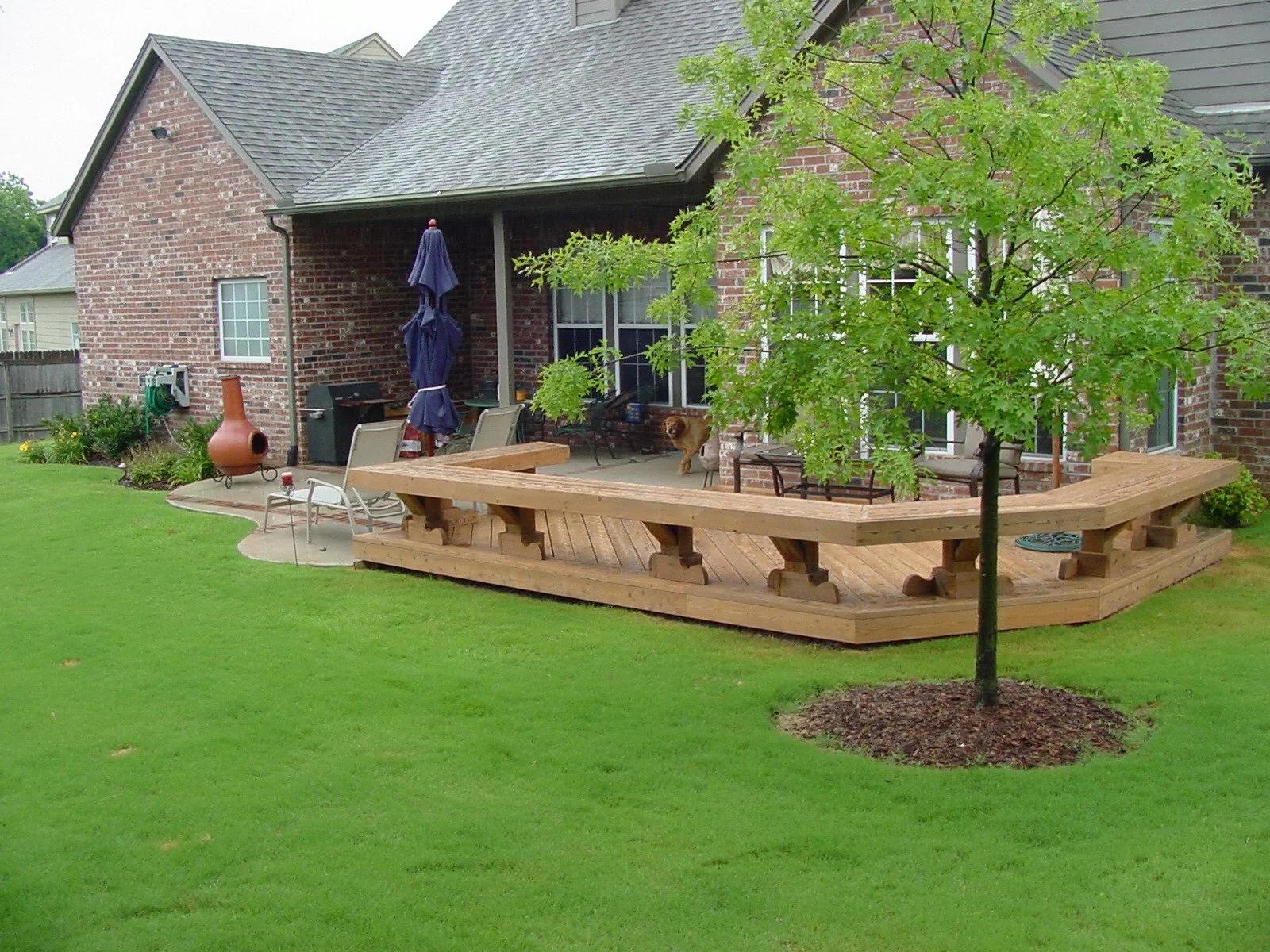 Backyard with wooden deck, green grass, small tree, house with brick exterior, patio furniture, umbrella, firepit, and a dog.