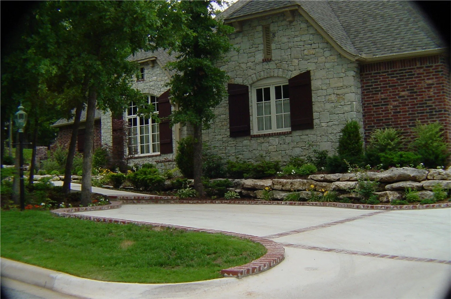 Front view of a house with stone and brick exterior, surrounded by greenery, shrubs, and trees, with a concrete driveway and brick-edged landscaping.