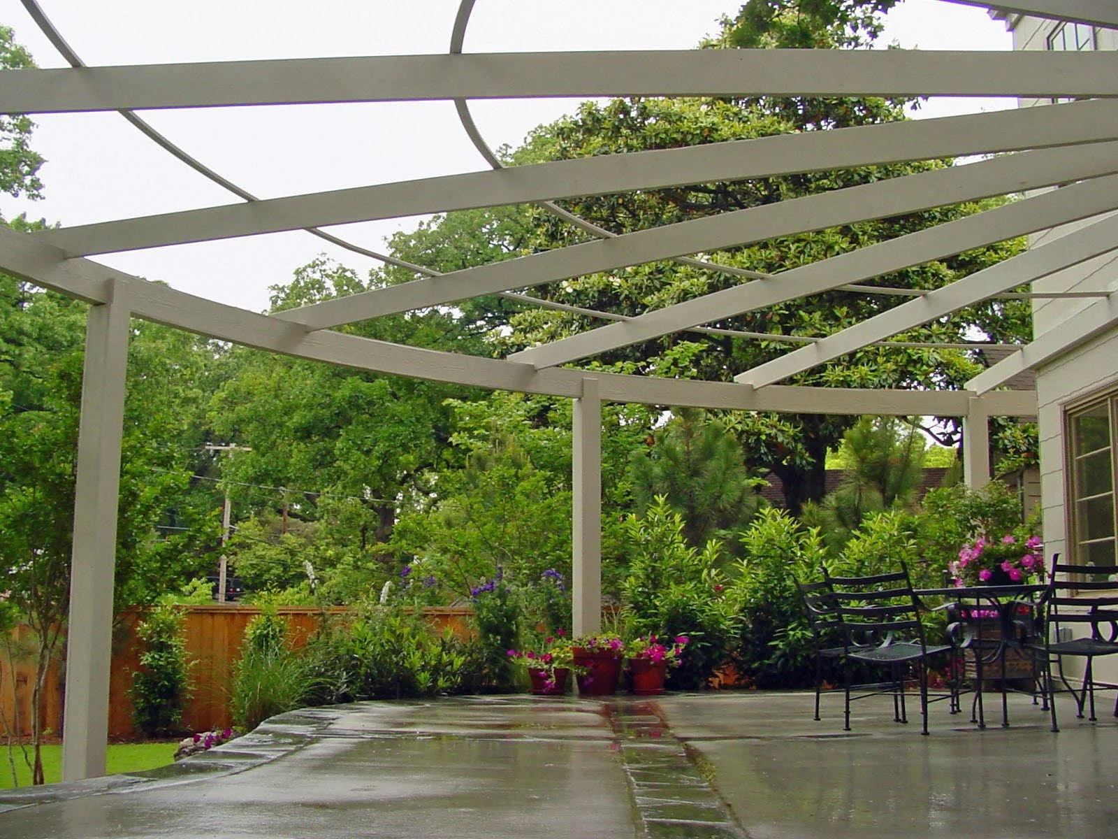 A backyard patio with a white pergola frame, wet concrete floor, green trees, potted plants, and outdoor furniture.