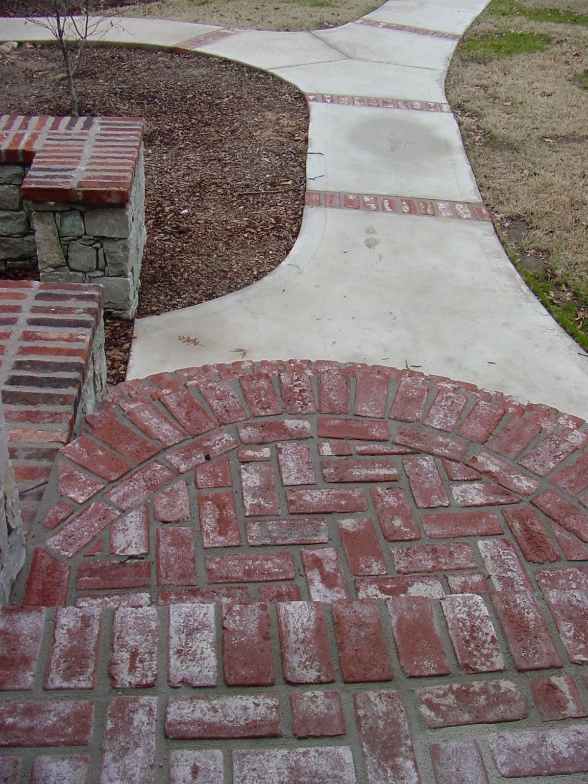 Concrete walkway with brick accents, curved design, and adjacent soil and grass areas.